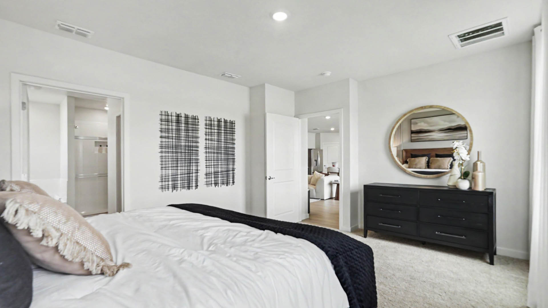 A modern bedroom features a neatly made bed with textured pillows and a dark throw, a black dresser with decorative items, a round mirror above it, and two abstract wall art pieces near an open doorway.