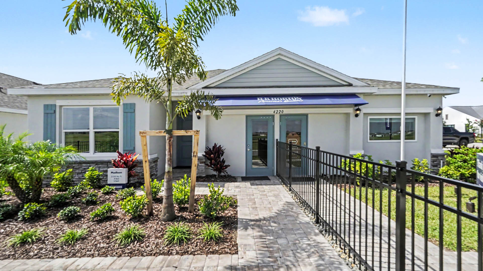 A single-story building with a paved walkway, a ramp with black railings, and landscaping featuring palm trees and shrubs under a clear blue sky.