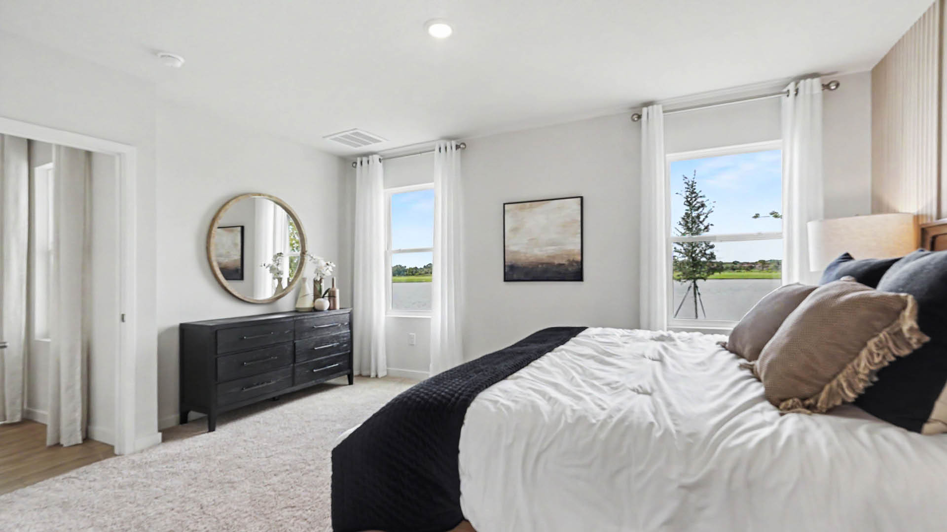 A modern bedroom features a neatly made bed with white and dark bedding, a black dresser with decorative items, a round mirror, two windows with white curtains, and a neutral color palette.