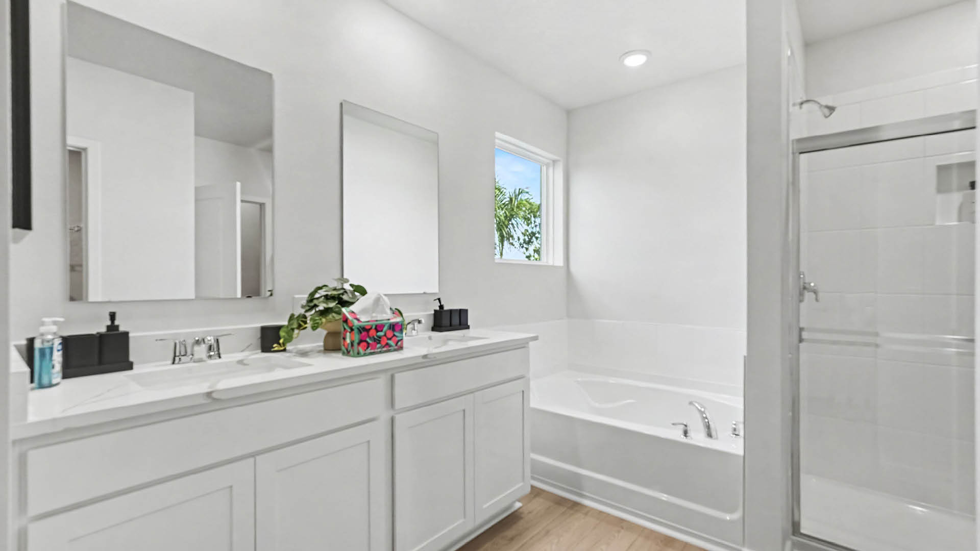 A modern white bathroom features a double sink vanity with large mirrors, a built-in bathtub beneath a small window, and a glass-enclosed shower.