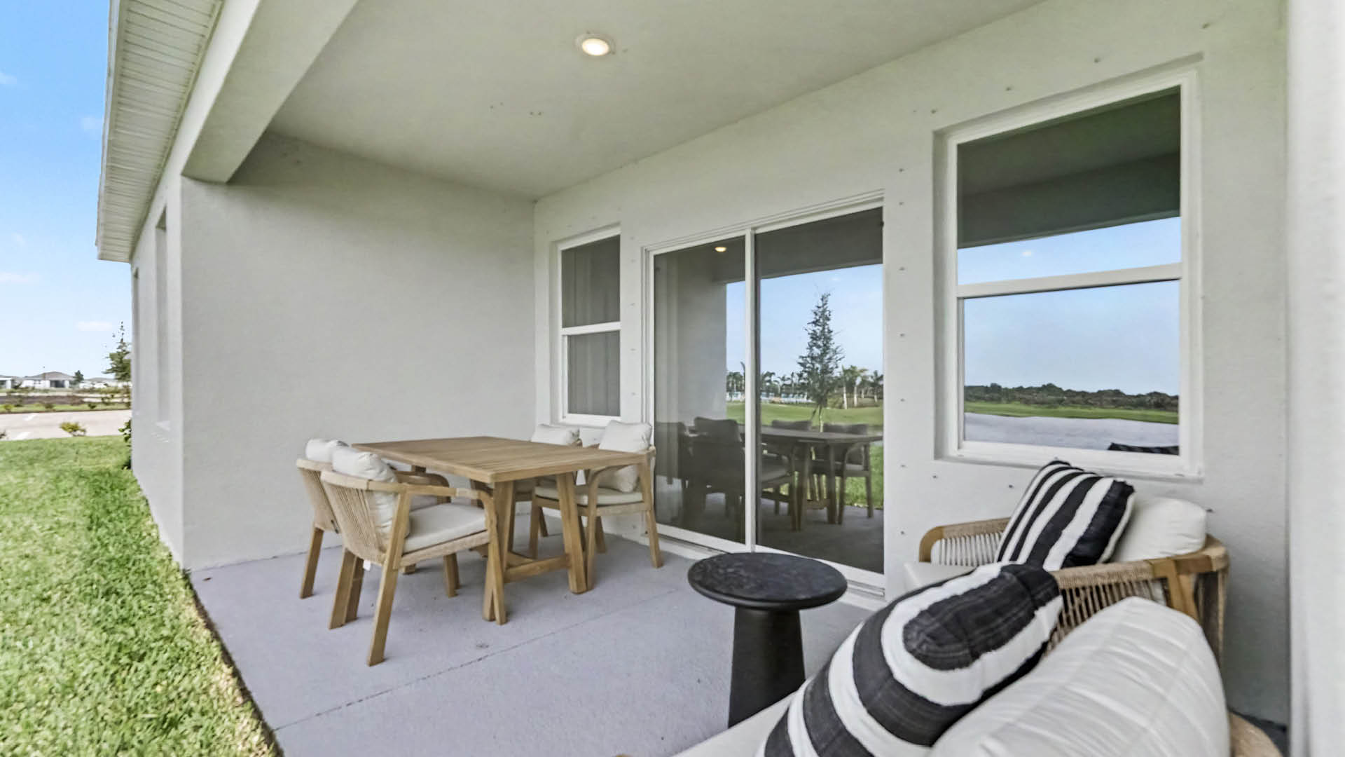 A covered outdoor patio features a wooden dining table with four chairs and a seating area with cushioned wicker furniture and striped pillows overlooking a grassy yard.