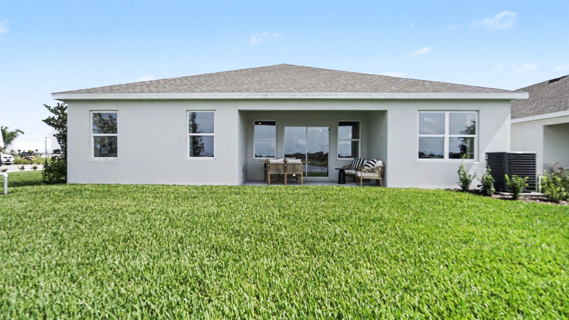 A modern single-story house with a large green lawn, a covered patio with outdoor furniture, and clear blue sky in the background.