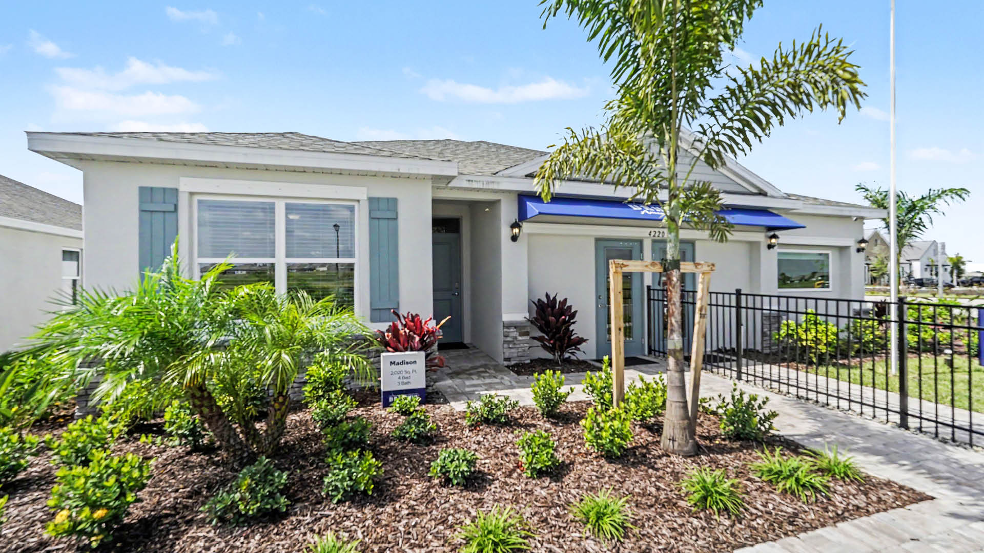 A single-story house with light blue shutters, a small front porch, and a landscaped garden with various plants and a palm tree under a clear blue sky.