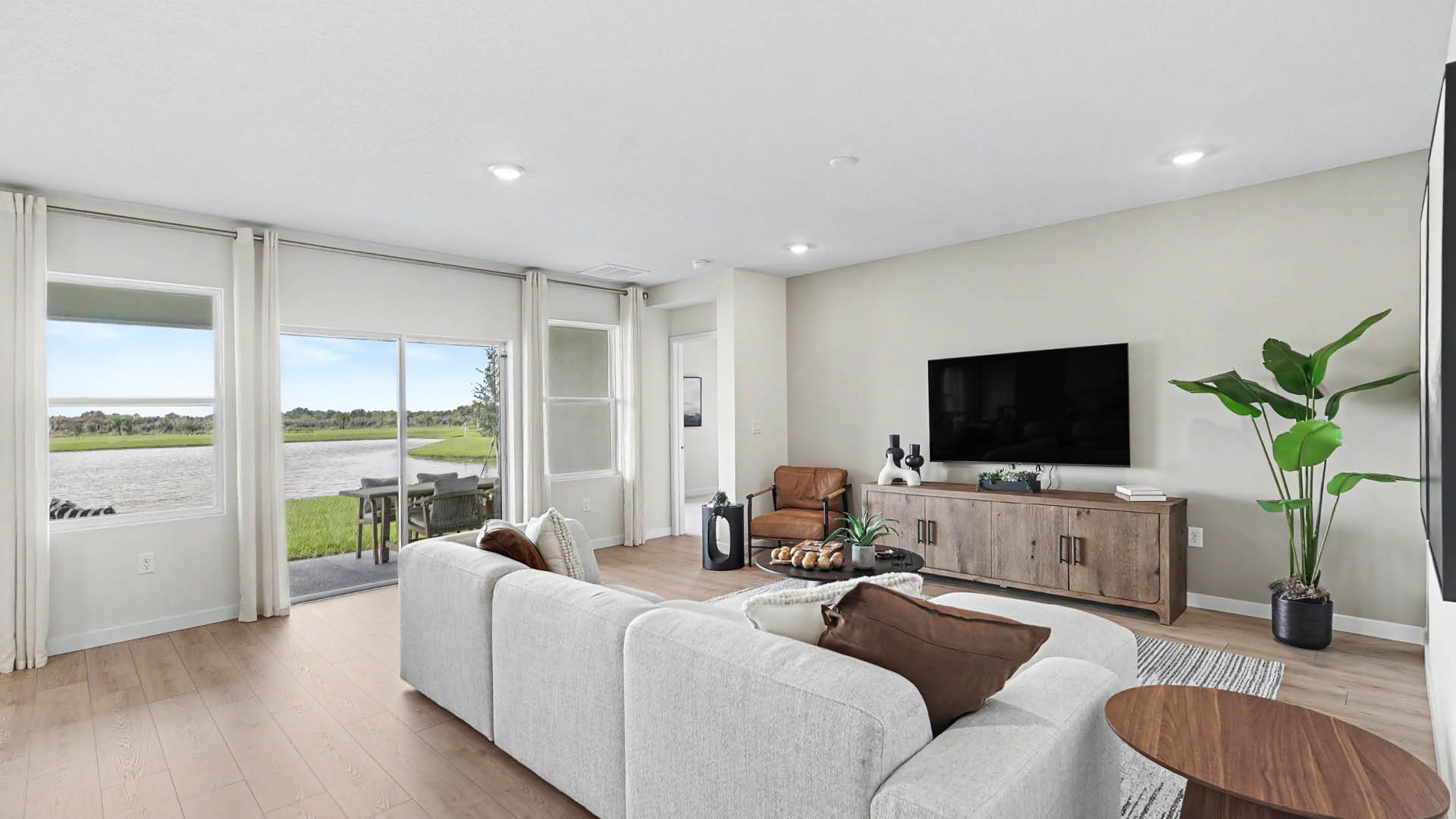 A modern living room features a large light gray sectional sofa with brown pillows, a wooden coffee table, a flat-screen TV mounted above a wooden console, a potted plant, and sliding glass doors opening to a covered patio with a view of a lake and greenery.