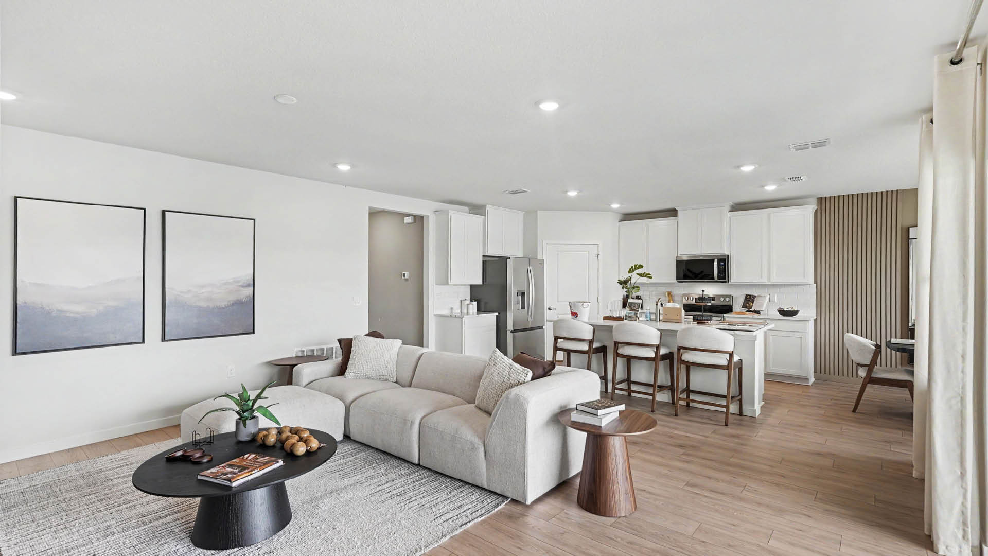 A modern open-concept living room and kitchen features a light gray sectional sofa, two framed landscape prints on the wall, a round wooden side table, a black coffee table with books and decor, and a kitchen island with three bar stools.