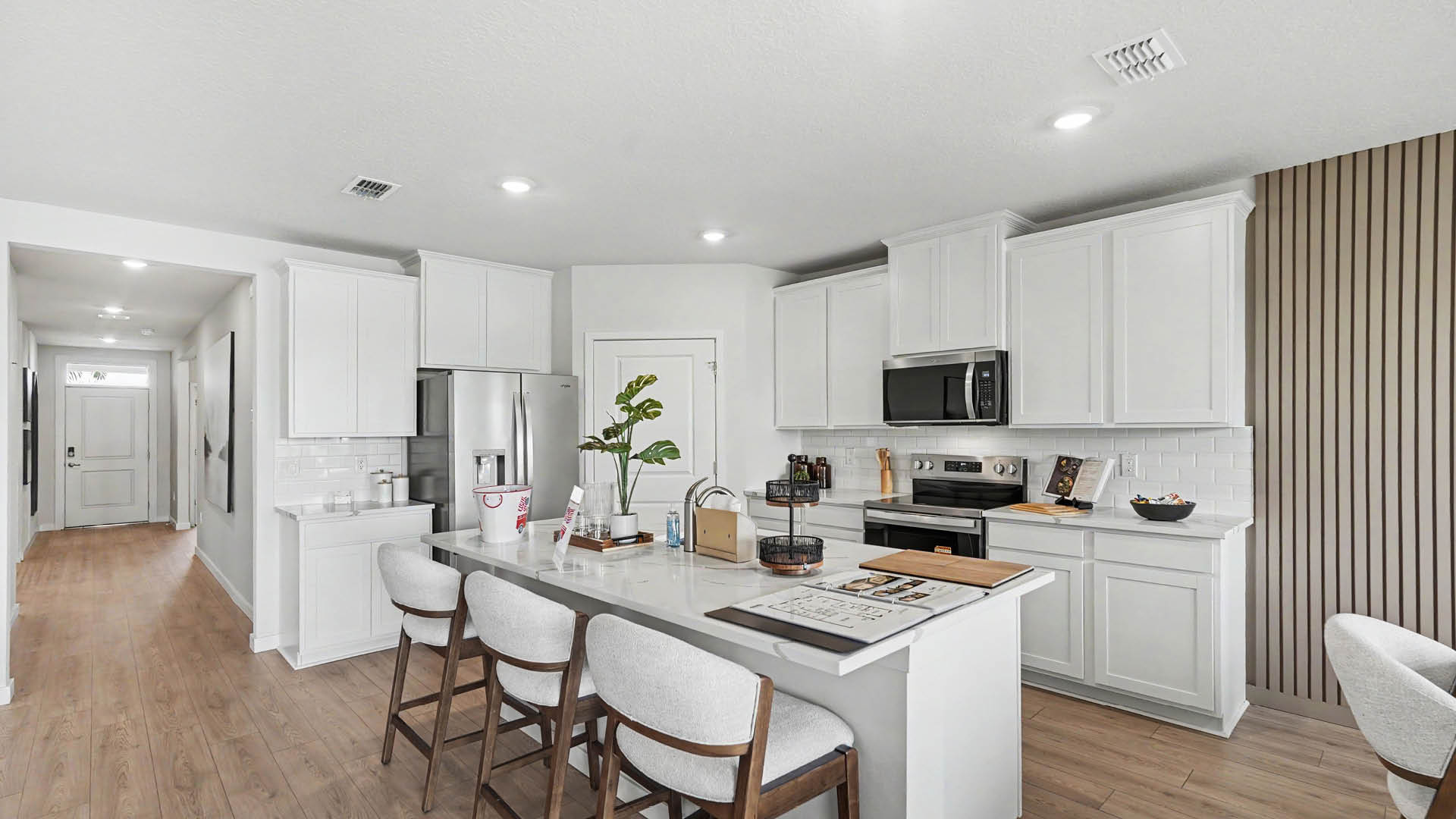 A modern kitchen features white cabinetry, a large island with three cushioned stools, stainless steel appliances, and Mohawk ® Revwood Select Plank Flooring extending into a hallway.