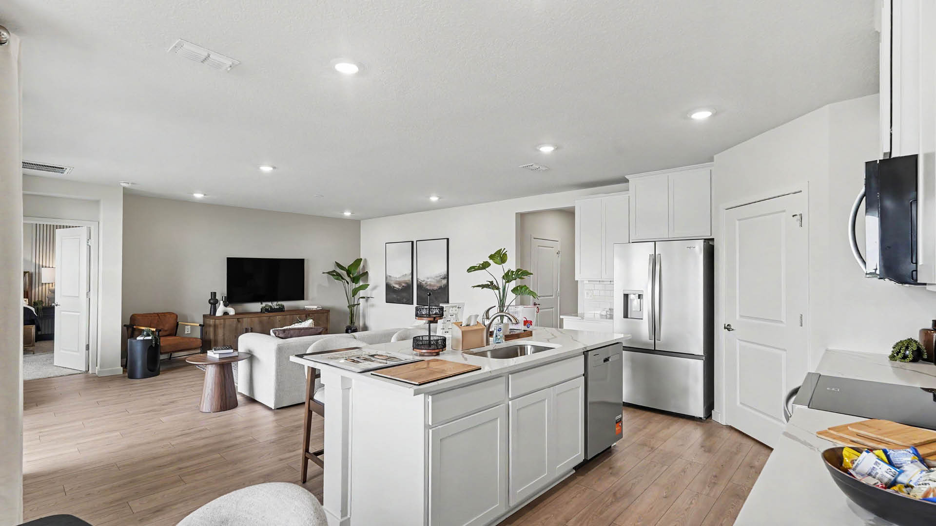 A modern open-concept kitchen and living area features white cabinetry, stainless steel appliances, a kitchen island with a sink and cutting board, and a seating area with a television and plants.