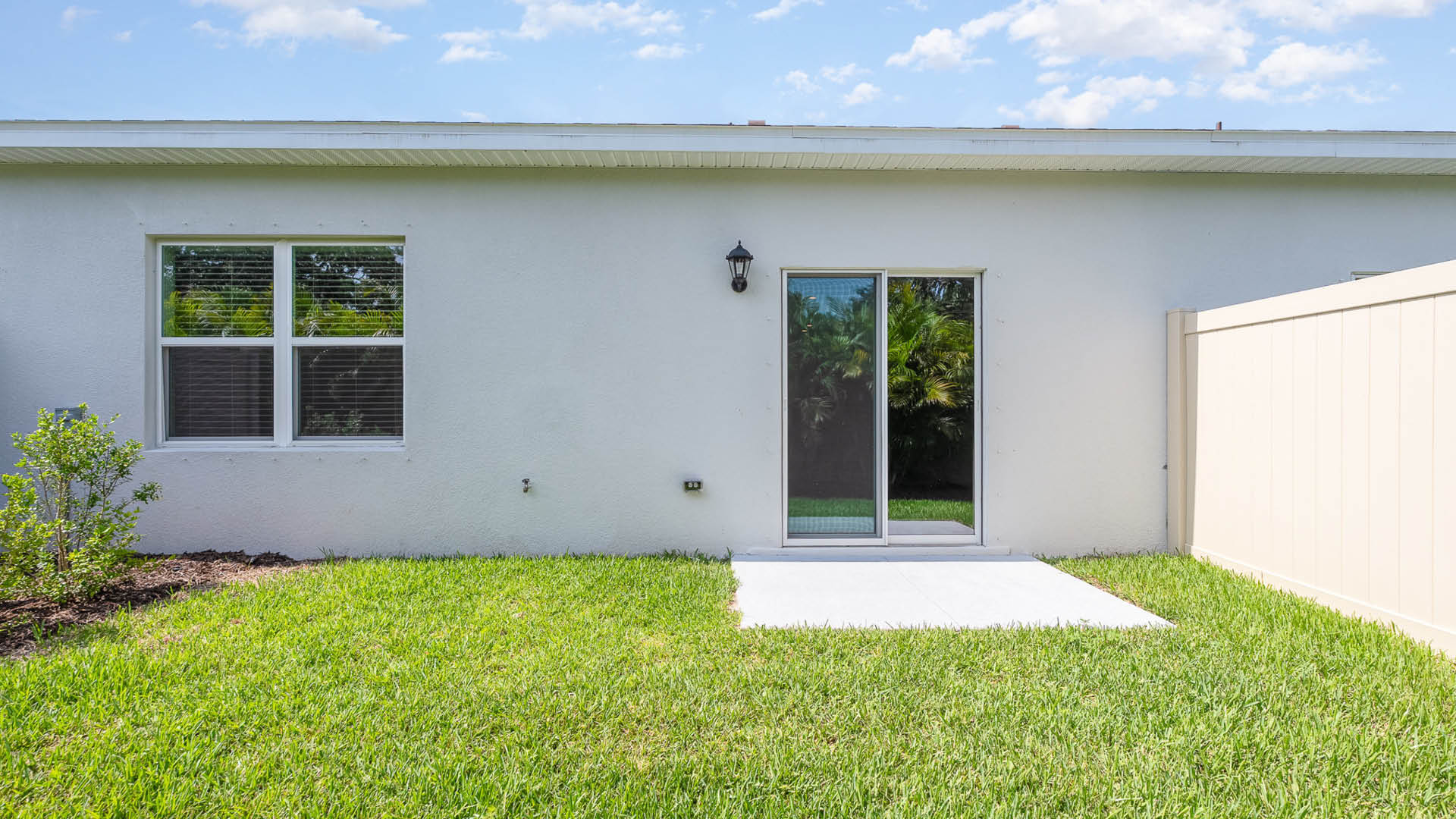 21. Rear view of a house featuring a white stucco exterior, sliding glass door, and a well-kept lawn with a small shrub.