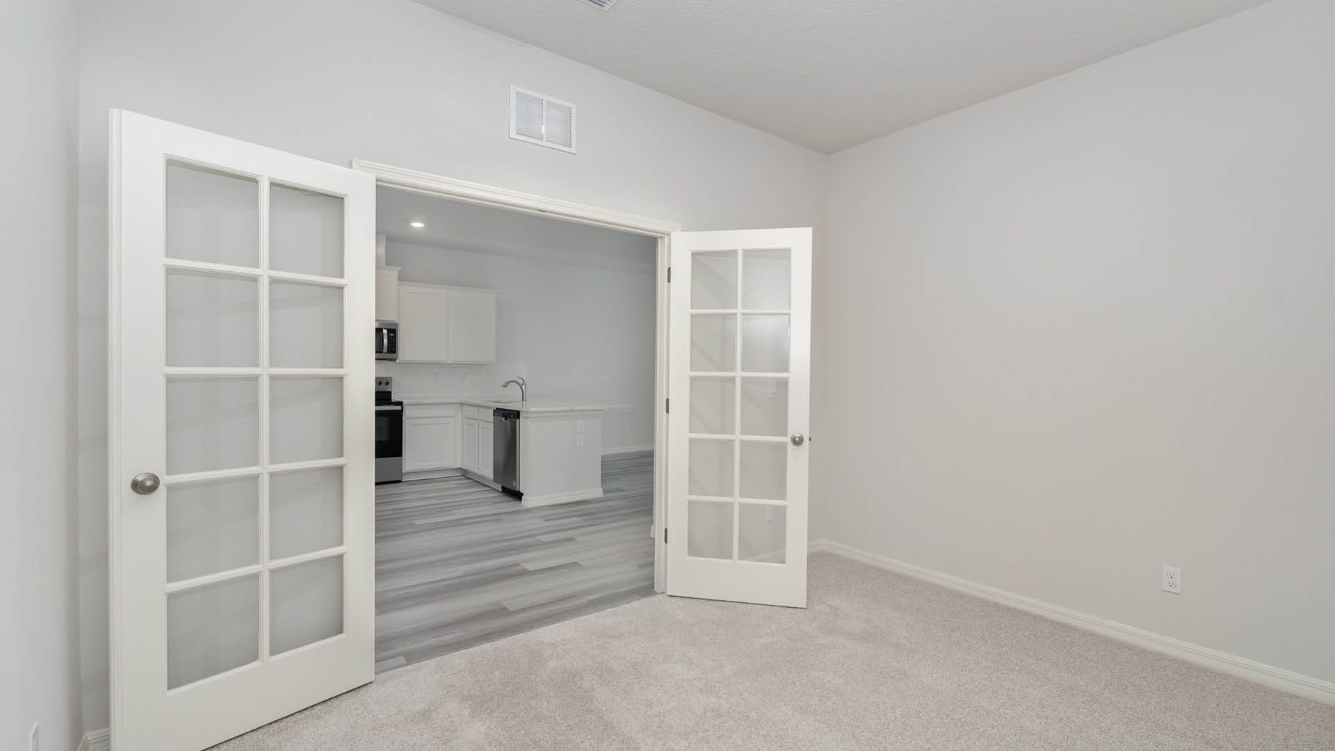 17. Bright room with carpeted floor, showcasing double glass doors leading to a modern kitchen with white cabinets and stainless appliances.