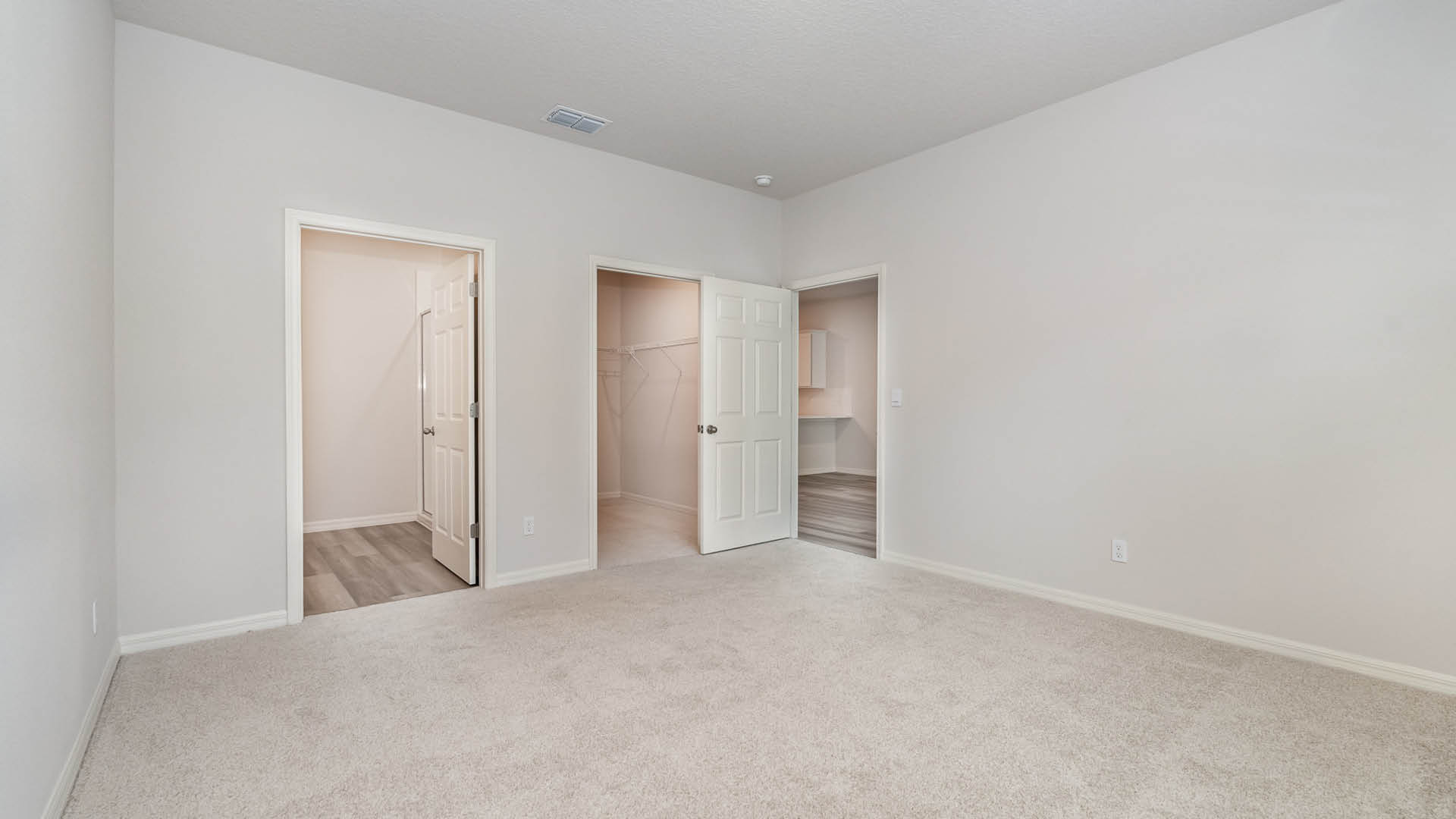 A large bedroom with beige carpet and white walls. Three open doors are visible. One is the entry to the bedroom, one is a large walk-in closet, and the other leads to an ensuite bathroom.