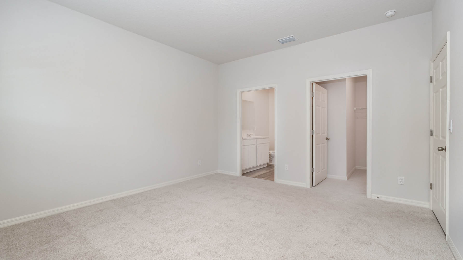A large bedroom with beige carpet and white walls. Two open doors are visible. One is a large walk-in closet and the other leads to an ensuite bathroom.