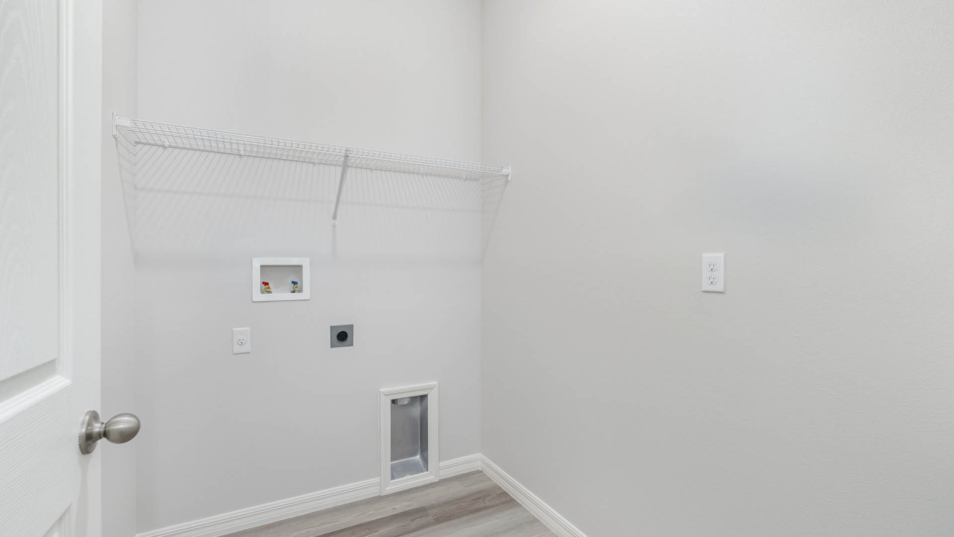 A laundry room with utility connections on the wall and white wire shelves. The open door to the room is visible.