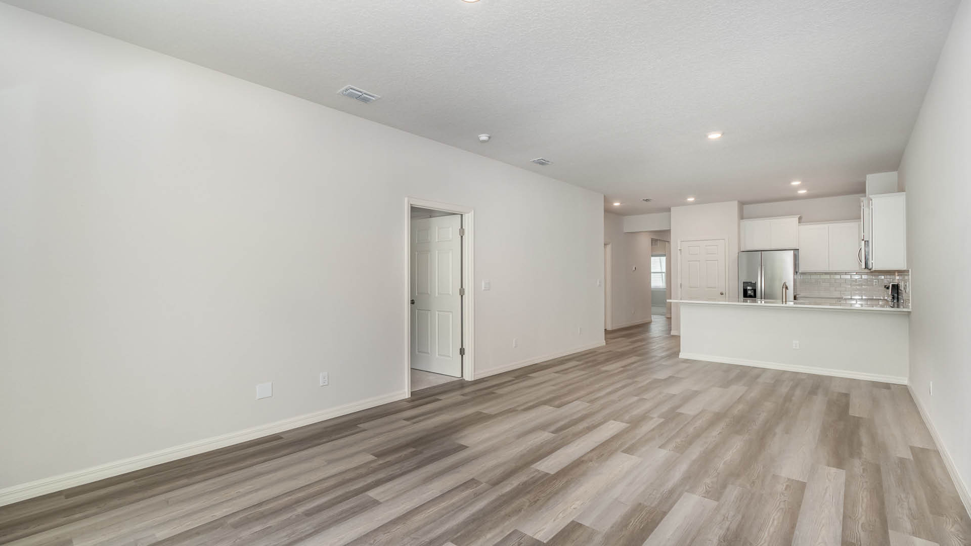 A bright, empty living and dining area with a view of a kitchen area. Several open doors leading to other areas are visible.