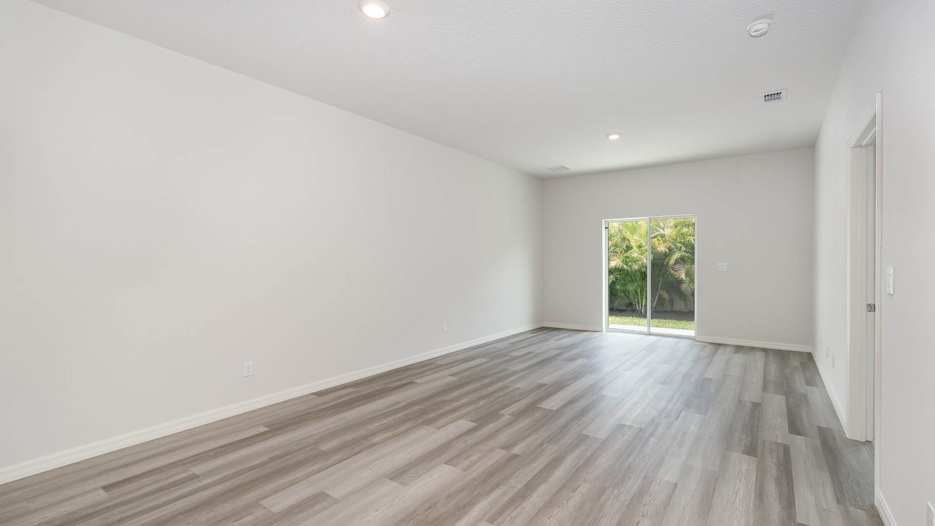 A bright, empty living and dining area featuring sliding glass doors leading to an outdoor patio. An open door leading to a bedroom is visible.
