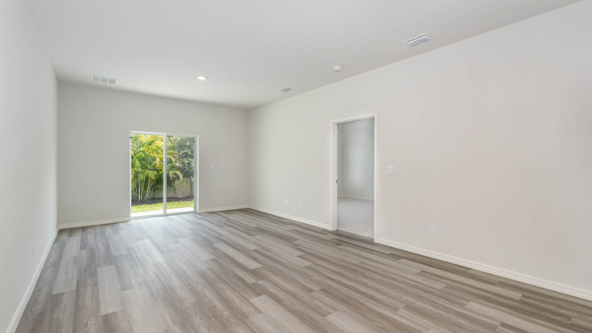 Bright, empty living room with Mohawk® Revwood Select Plank Flooring, large sliding glass doors to a garden, and an adjacent open door to another room.