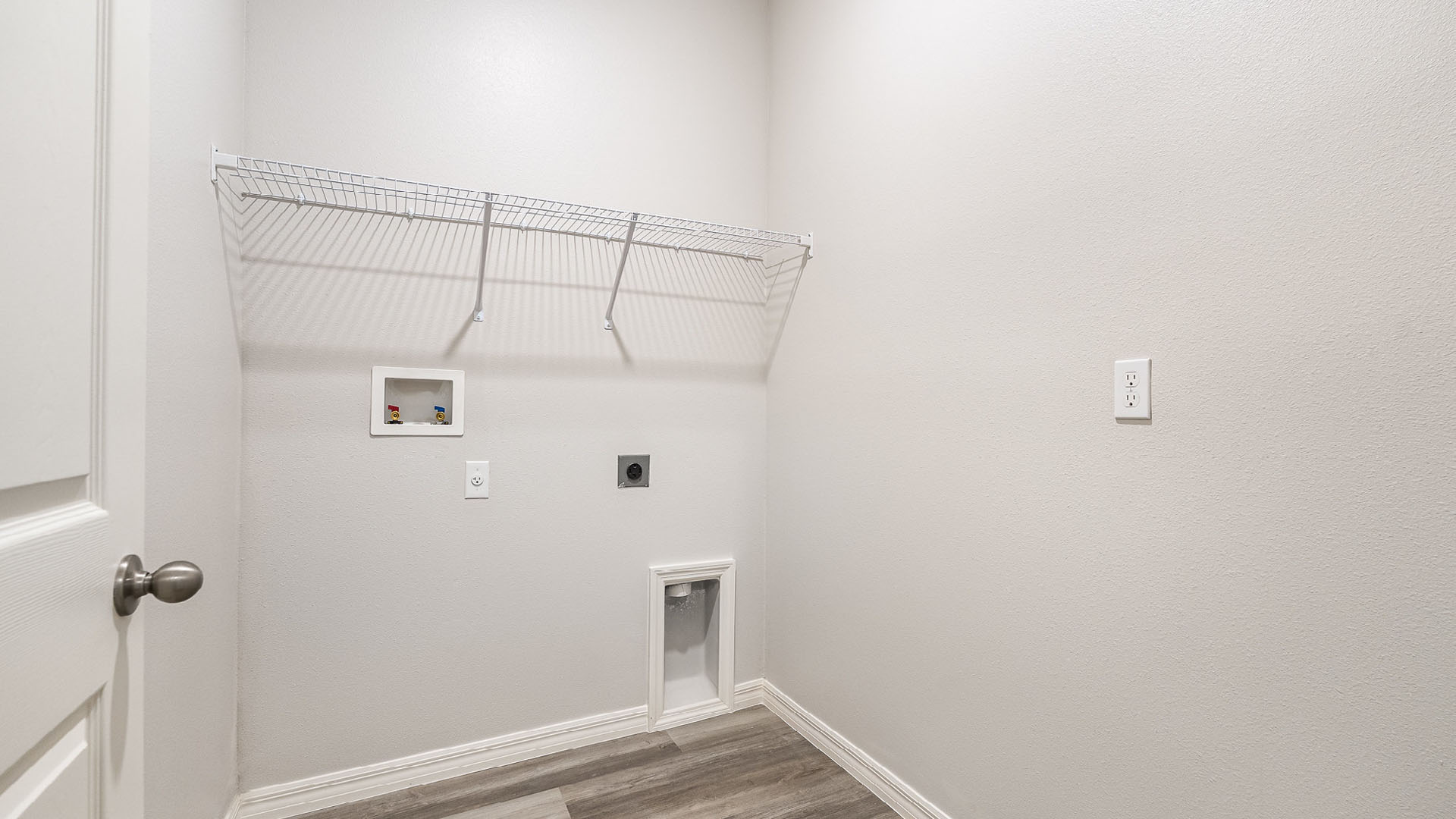 A laundry room with utility connections on the wall and white wire shelves. The open door to the room is visible.