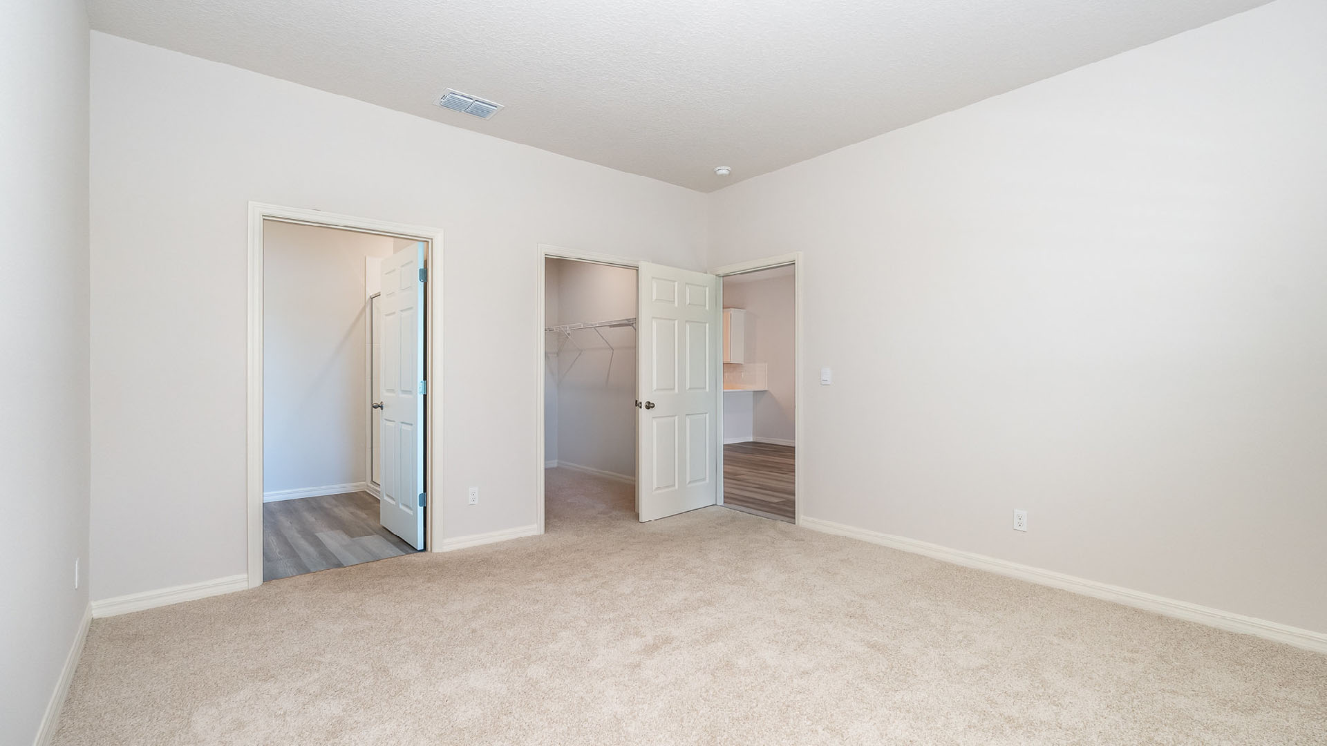 A large bedroom with beige carpet and white walls. Three open doors are visible. One is the entry to the bedroom, one is a large walk-in closet, and the other leads to an ensuite bathroom.