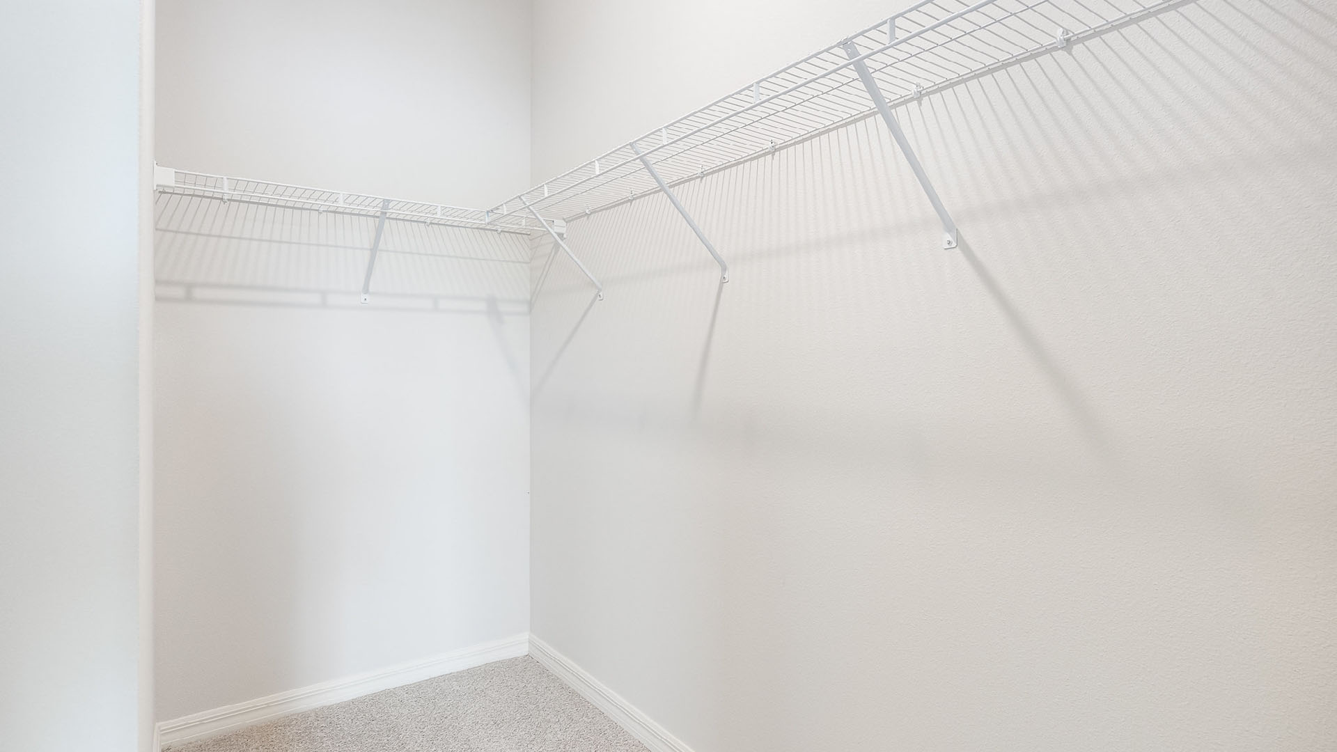 A spacious, walk-in closet with beige carpet, featuring white wire shelves against a light-colored wall.