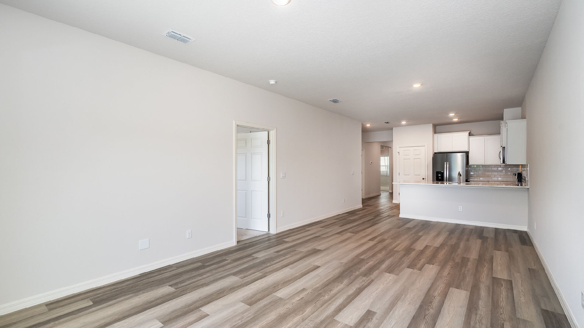 A bright, empty living and dining area with a view of a kitchen area. Several open doors leading to other areas are visible.
