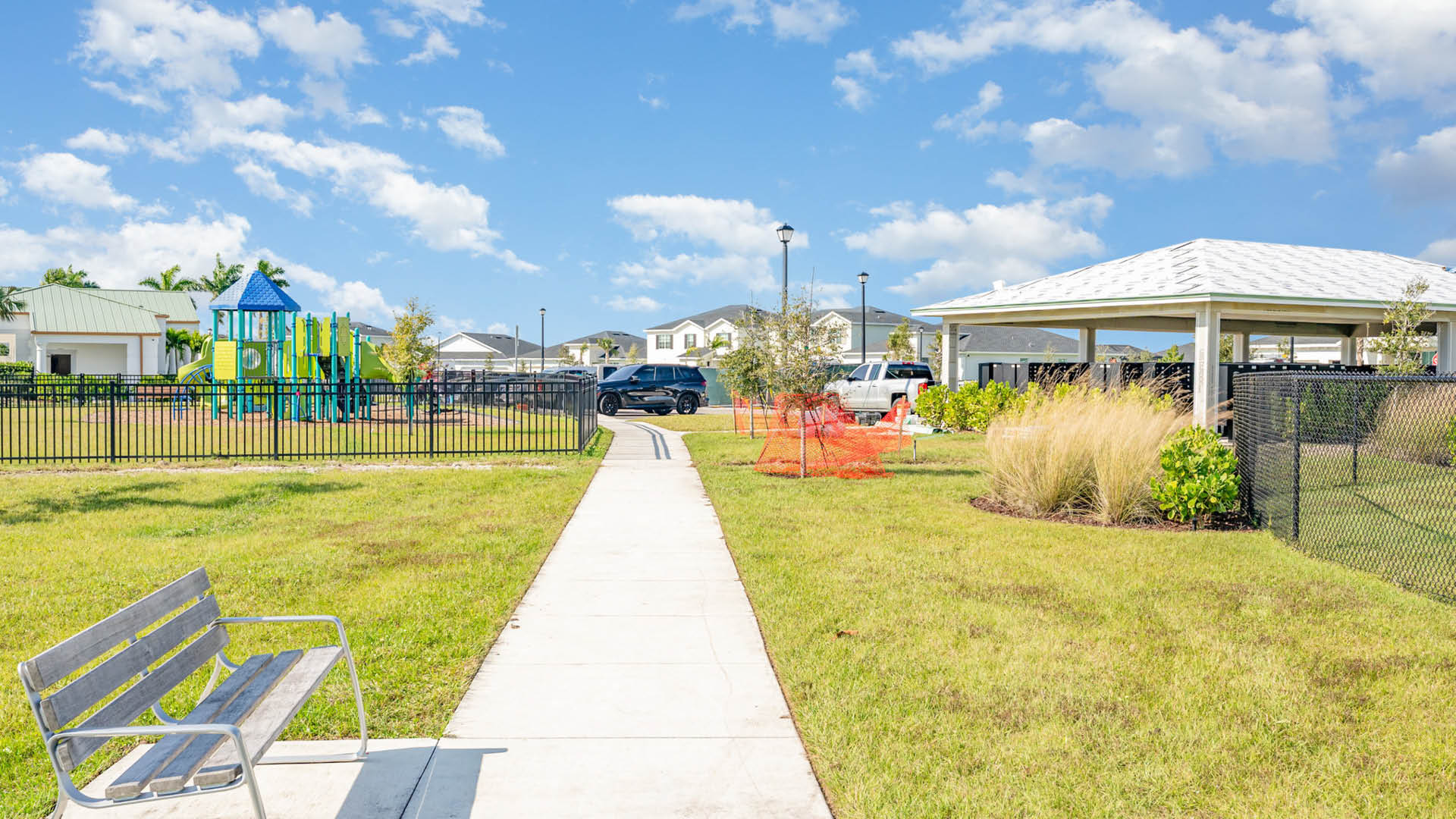 A sunny park with a pathway leading to a playground with colorful equipment on the left and a covered pavilion with parked cars on the right.