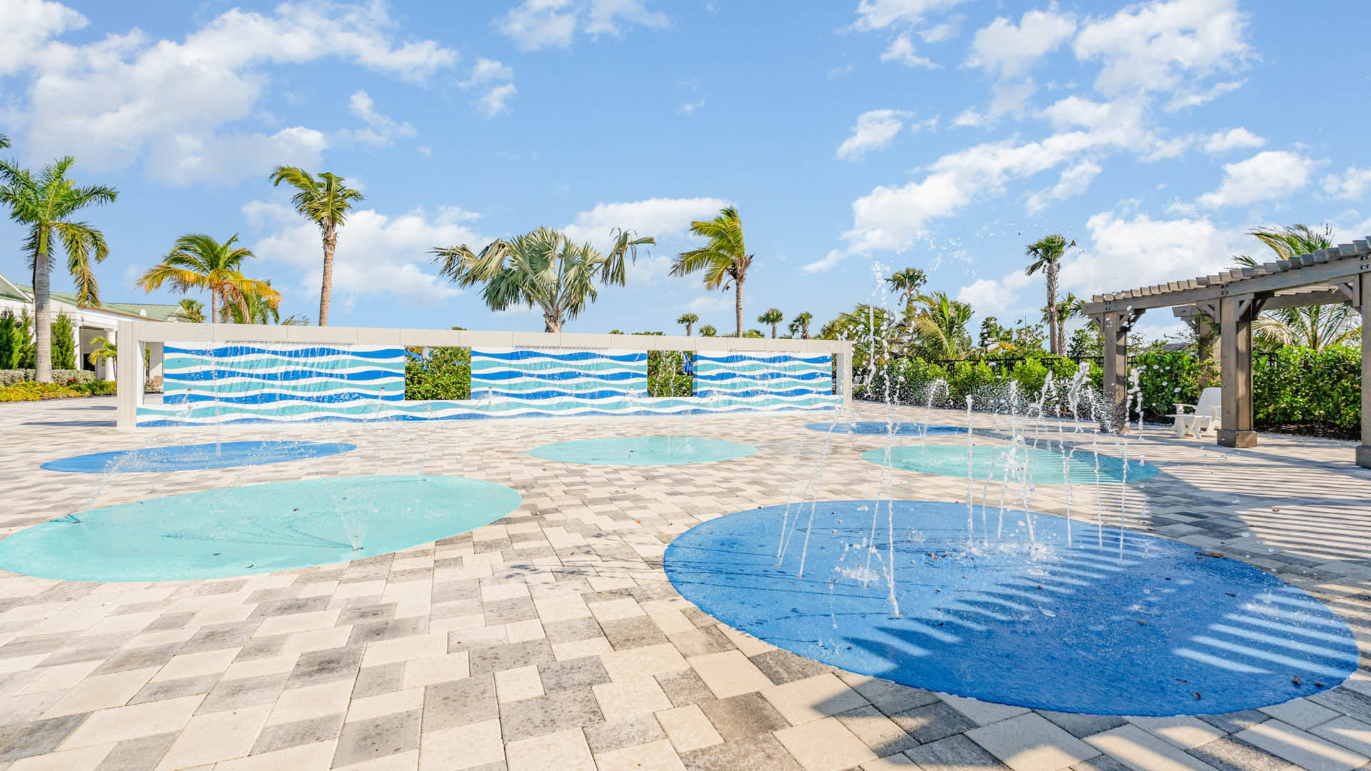 A splash pad with colorful blue and teal circles features water fountains. Palm trees surround the area, under a bright blue sky scattered with clouds.