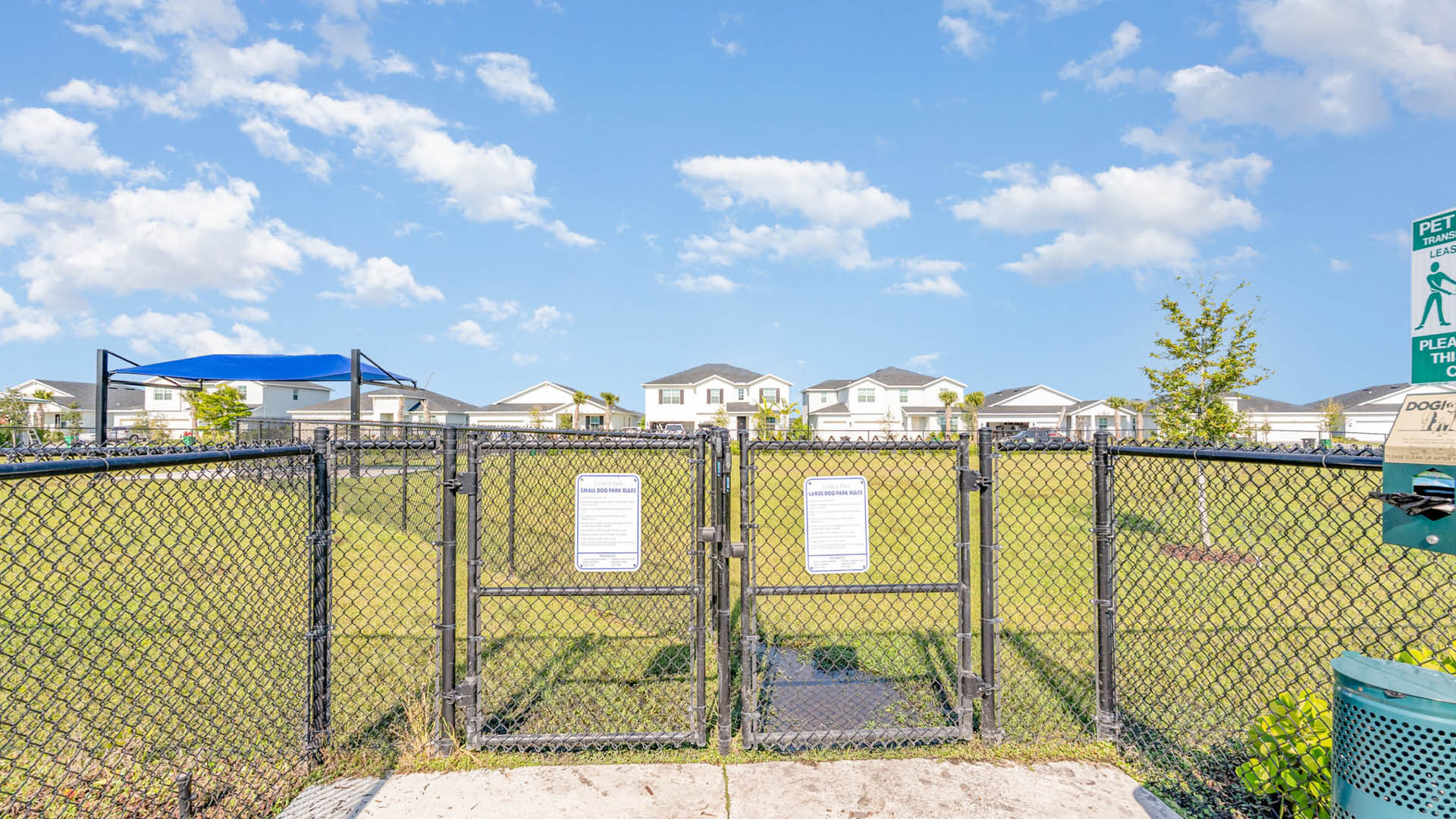 Chain-link fence with closed gate at dog park entrance. Lush green field and homes in background under a bright blue sky with clouds.