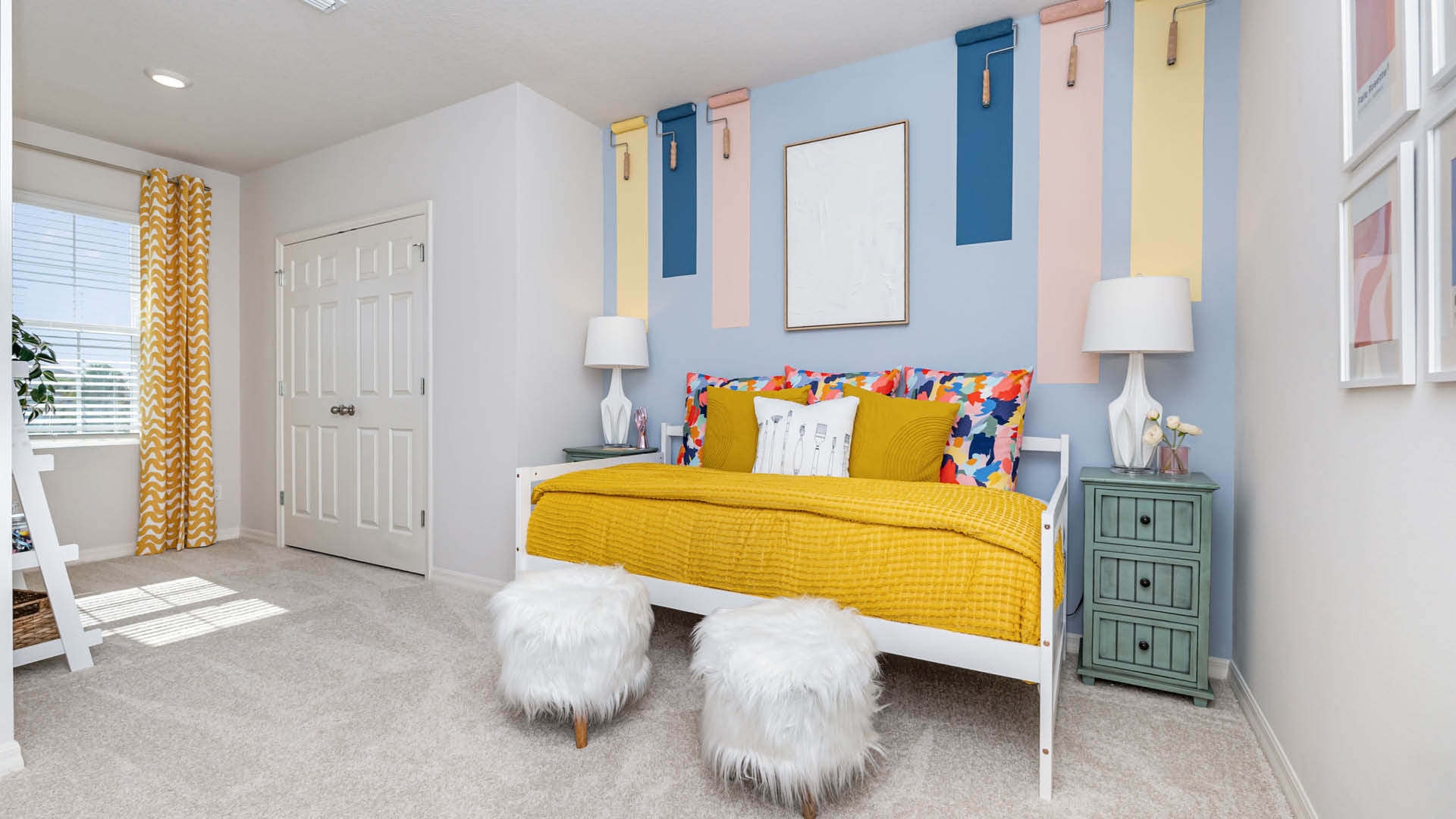 Bright bedroom with colorful accents. A yellow bedspread with floral pillows contrasts against a blue wall. Two white furry stools add a playful touch.