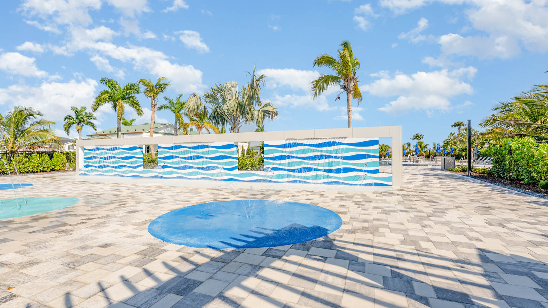 A sunny scene of a splash pad with palm trees, a wavy blue and white mural, clear blue sky, and decorative paving. It conveys a relaxed, tropical vibe.