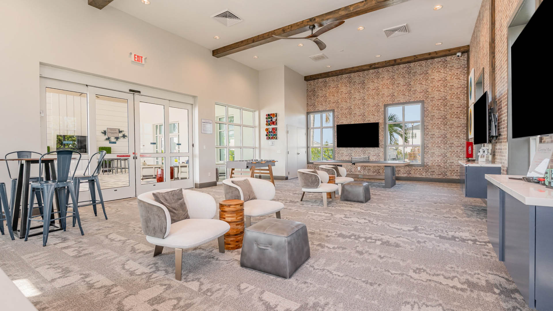 Modern lounge with a cozy atmosphere, featuring comfortable seating, a rustic brick wall with large windows, two flat-screen TVs, and high stools by the entrance.