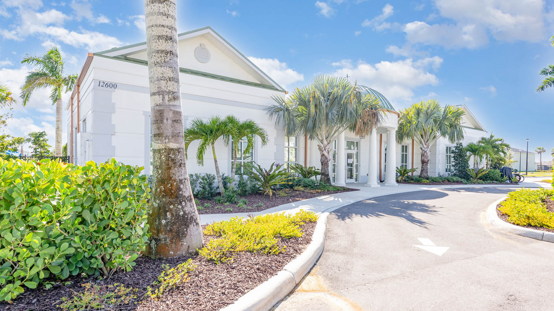 A bright, single-story white building with arched windows, surrounded by lush palm trees and greenery. A curved driveway leads to the entrance under a sunny, blue sky.
