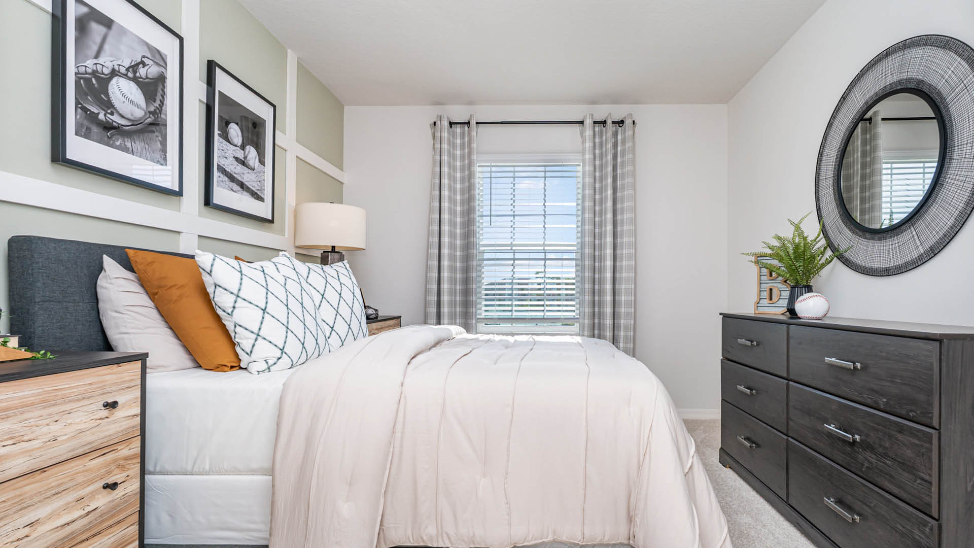 A neatly made bed with white and beige bedding sits beside a dresser and a window with gray curtains; black and white photos and a round mirror decorate the walls.