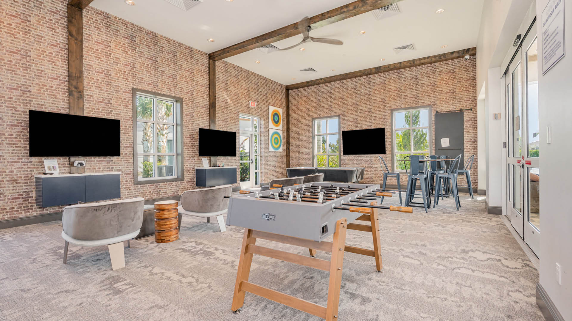Modern recreation room with exposed brick walls, large windows, three black flat-screen TVs, a foosball table, and cozy seating, exuding a relaxed ambiance.