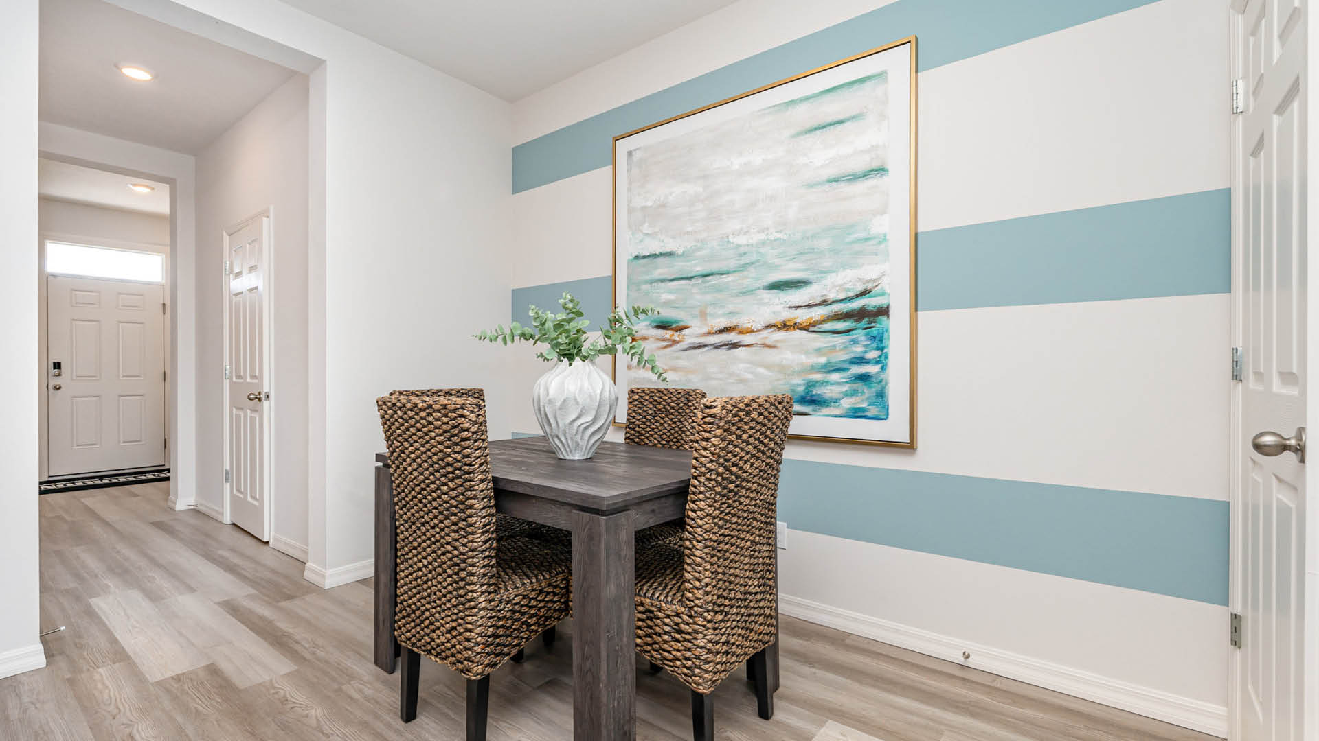 A cozy dining area featuring a wooden table, woven chairs, a vase with greenery, and a large abstract painting on striped walls.
