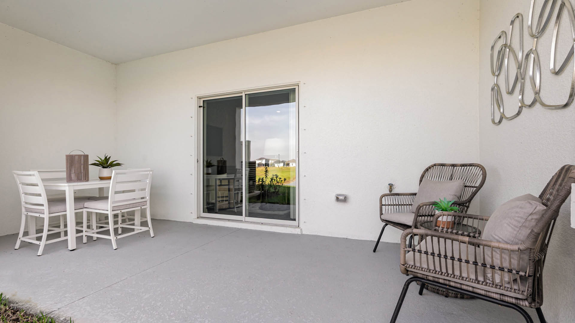 Covered patio with white walls featuring a white dining set on the left and two wicker chairs with cushions on the right. A glass door overlooks greenery.