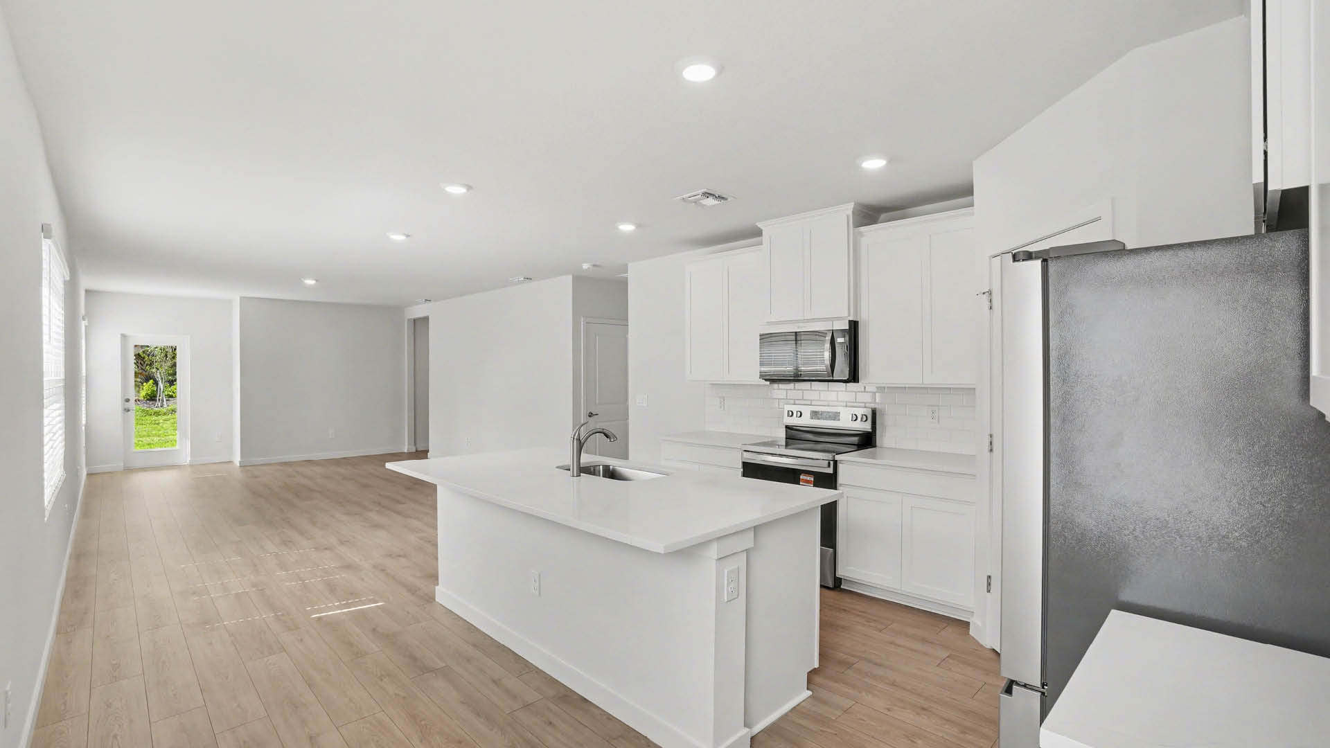 A modern kitchen with white cabinetry, quartz countertops, and stainless steel appliances. There is a view of a large dining and living area and the closed back door leading to a covered patio.