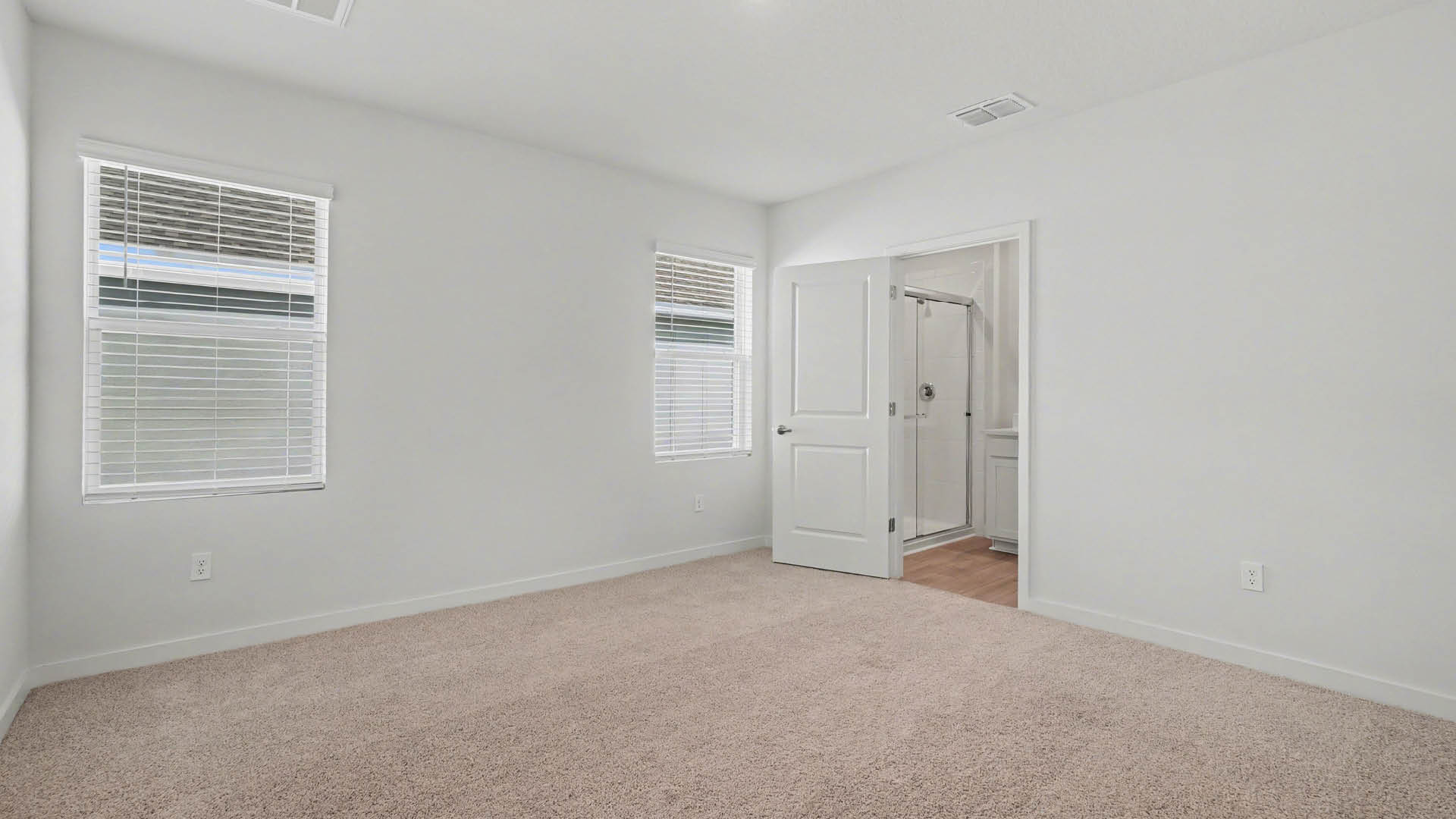 A large bedroom with beige carpet and two windows. An open door to an ensuite bathroom is visible.