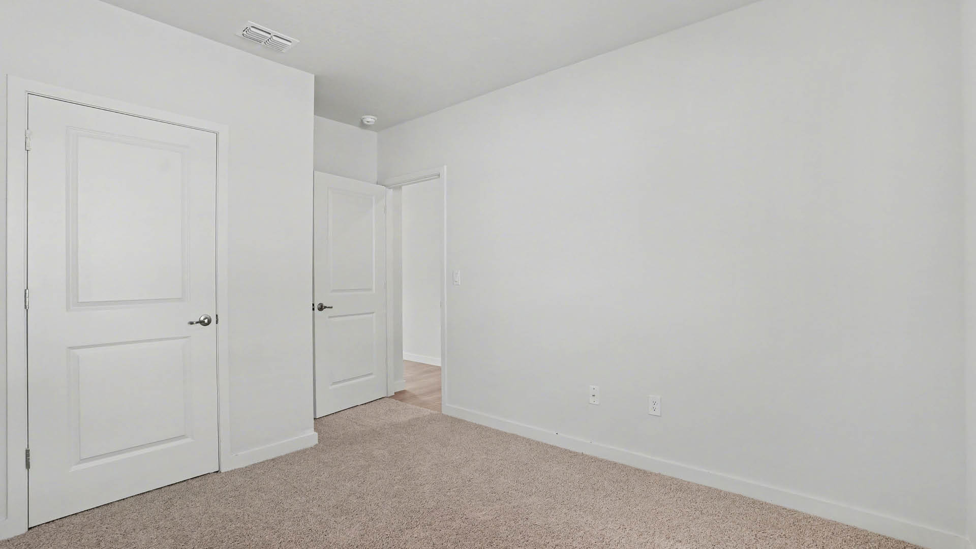 A large bedroom with beige carpet and white walls. An open door leading out of the room and a closed closet door are visible.