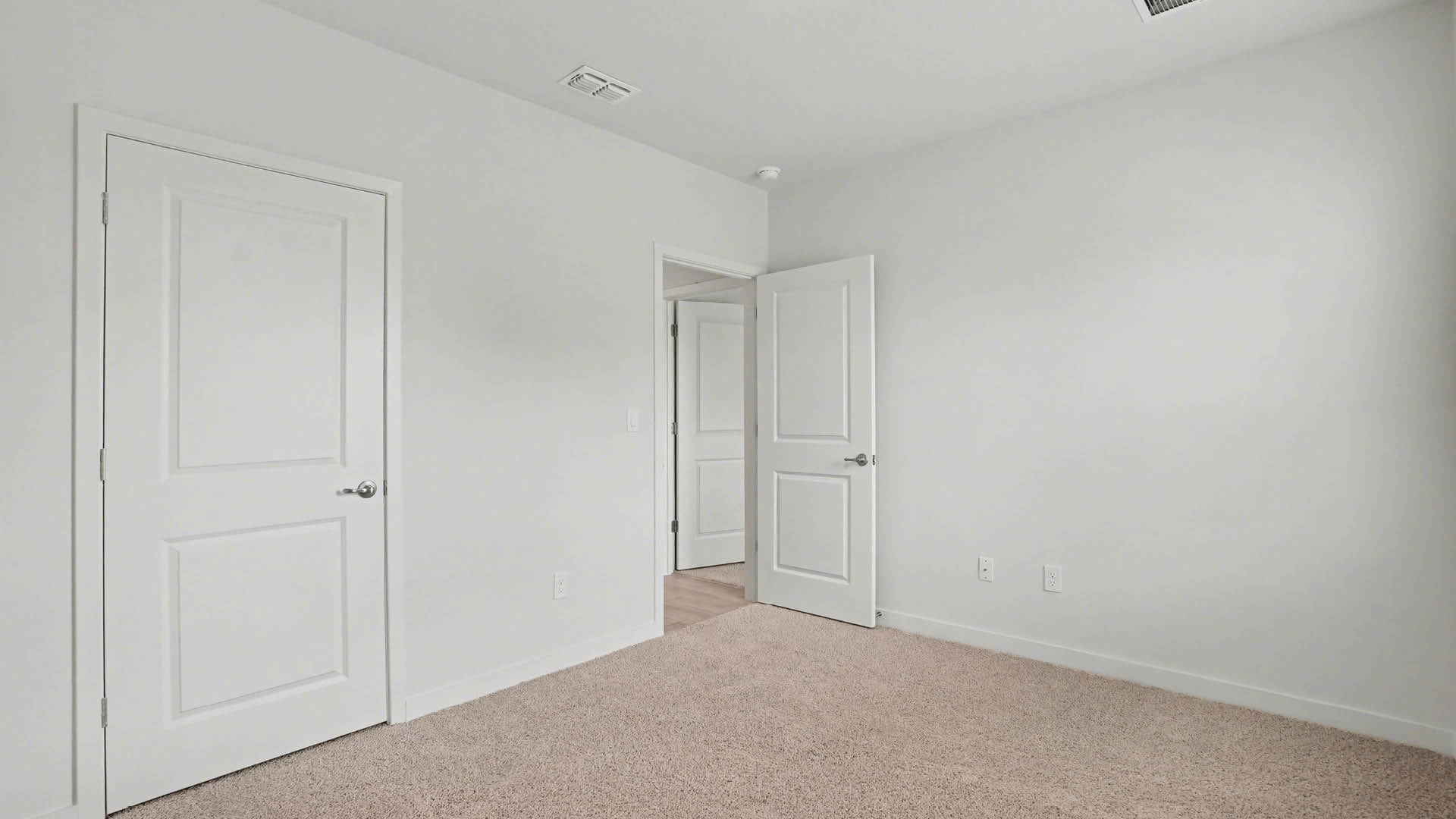 A large bedroom with beige carpet and white walls. An open door leading out of the room and a closed closet door are visible.
