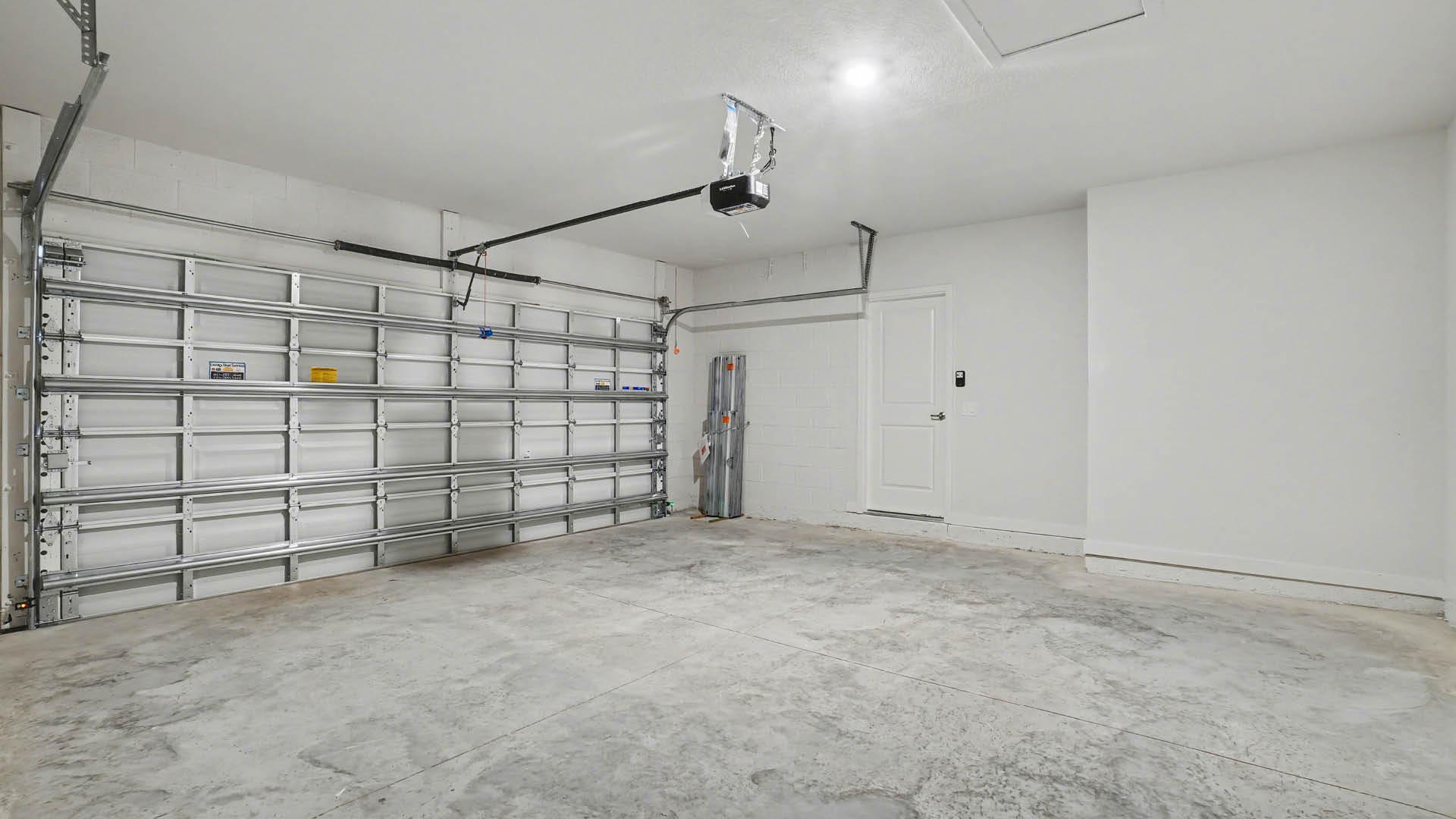 A spacious, 2-car garage with concrete flooring, a garage door, and a white cinder block wall. An access panel in the ceiling is visible.