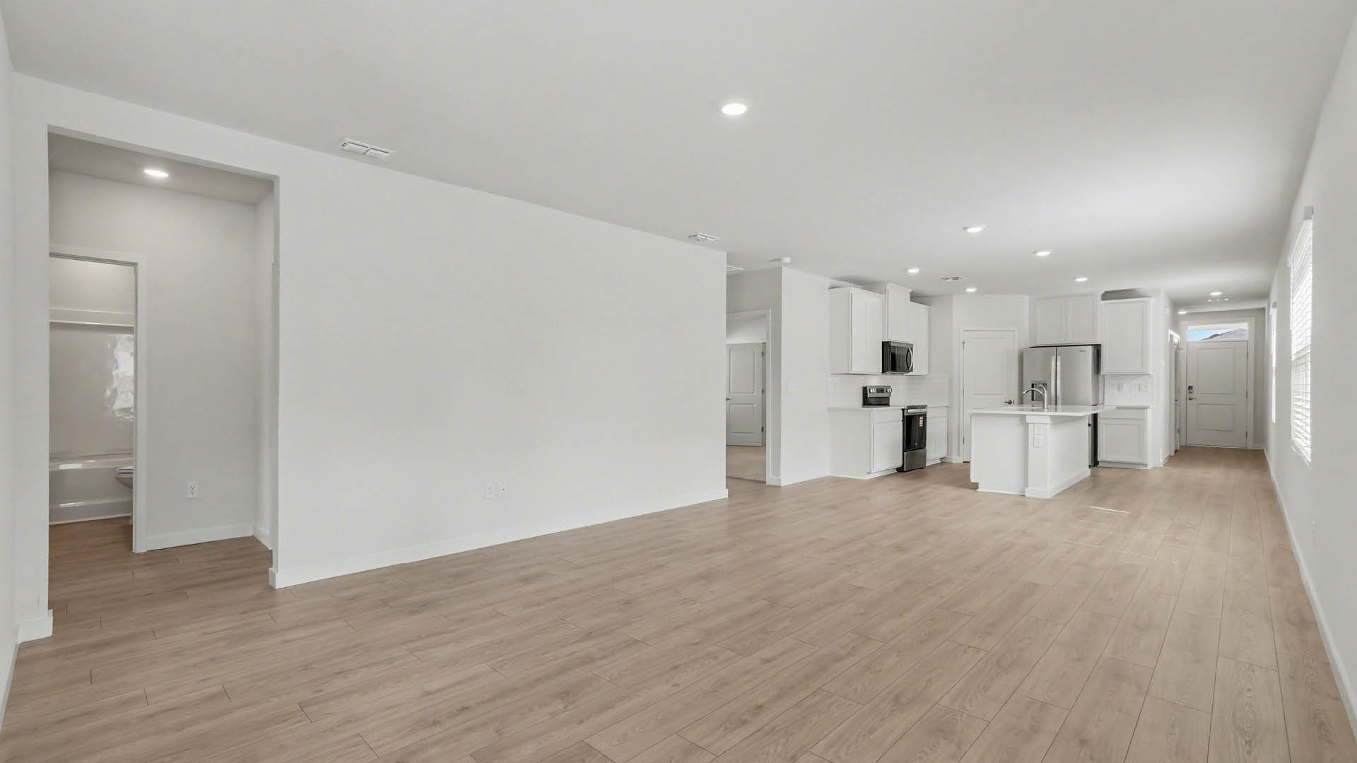 A spacious room, with a view of a kitchen area and Mohawk® Revwood Select Plank Flooring throughout. Also visible are the closed front door, a hallway leading to another area, and an open bathroom door.