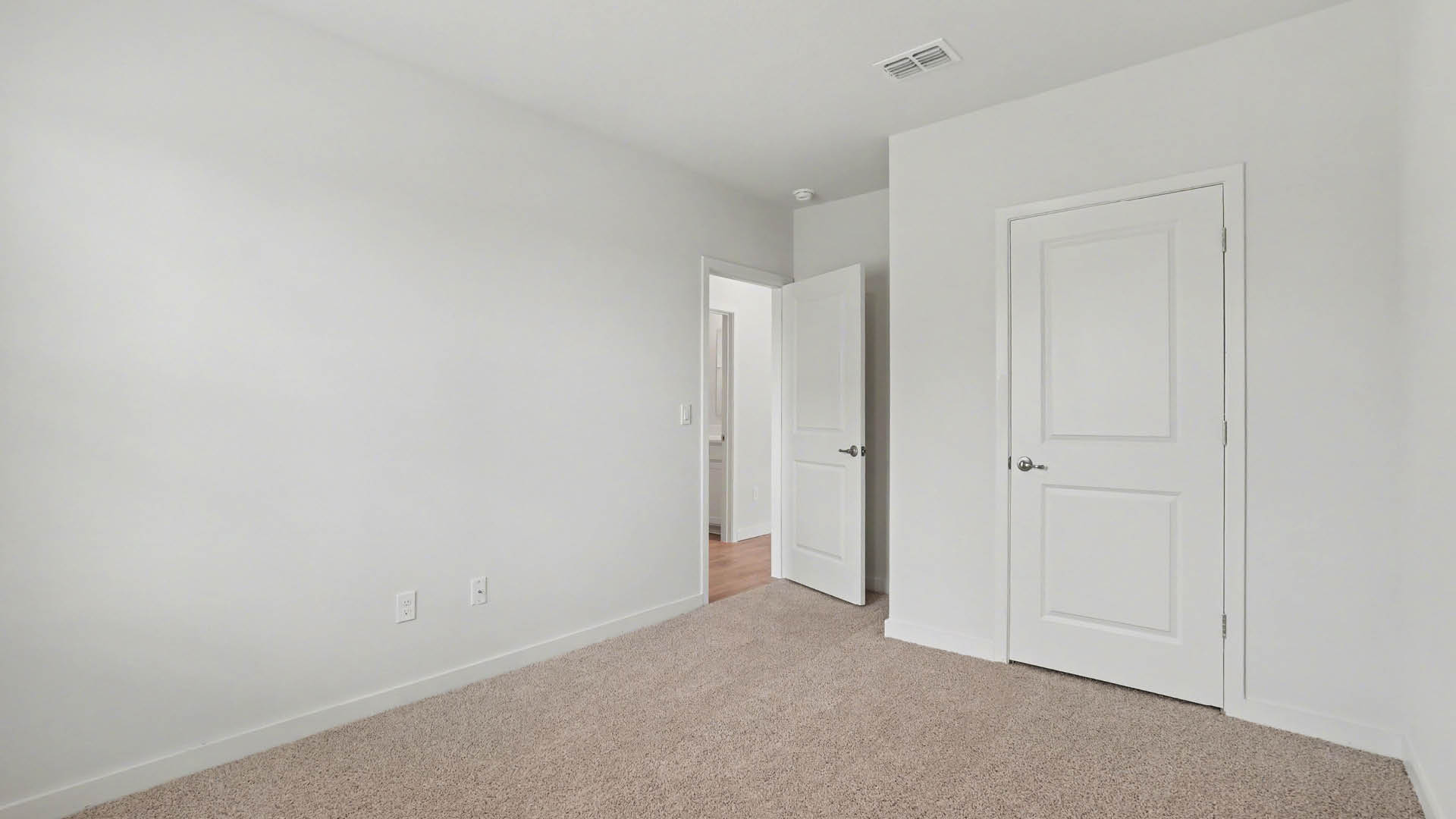 A large bedroom with beige carpet, white walls, and a large window with blinds, allowing natural light to illuminate the space.