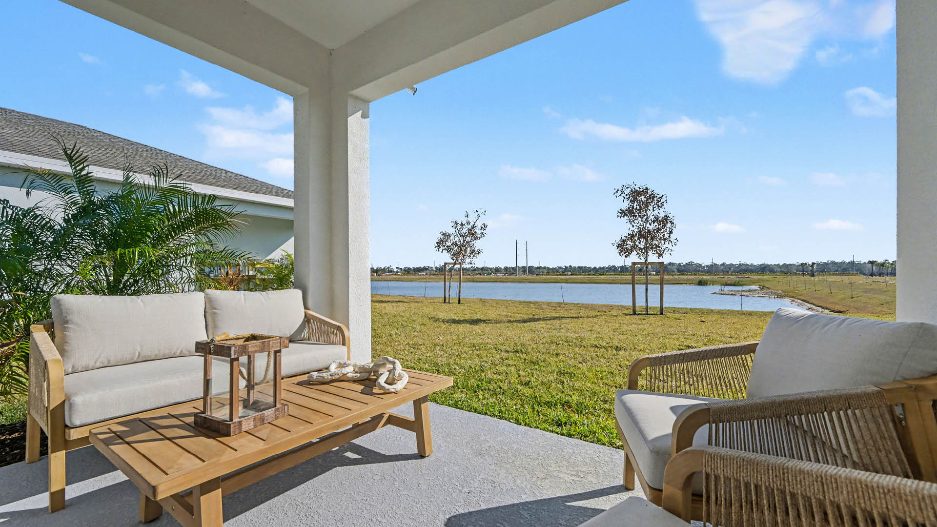 Seating area on a covered patio with a wooden table, surrounded by greenery, overlooking a tranquil lake.