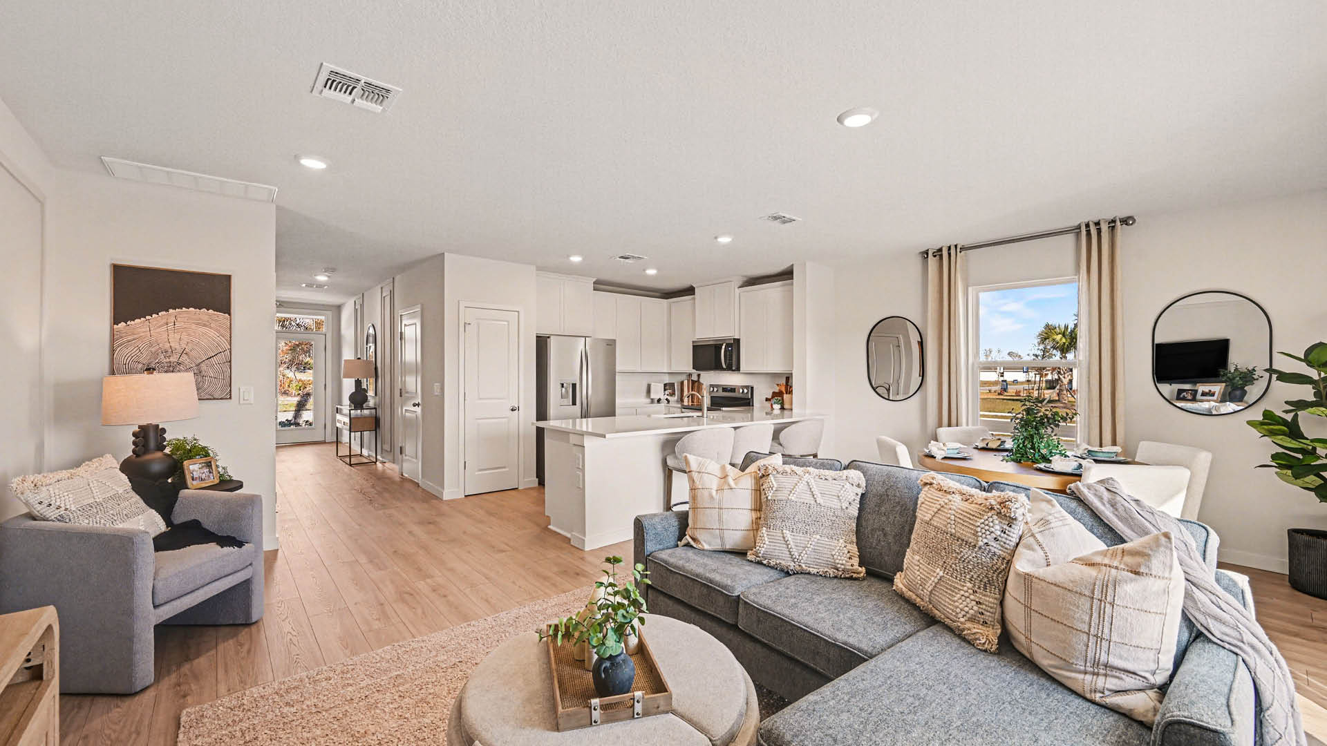 A spacious living room, leading into a dining and kitchen area with the entryway hallway and front door visible beyond. There is a large window and Mohawk® Revwood Select Plank Flooring throughout.