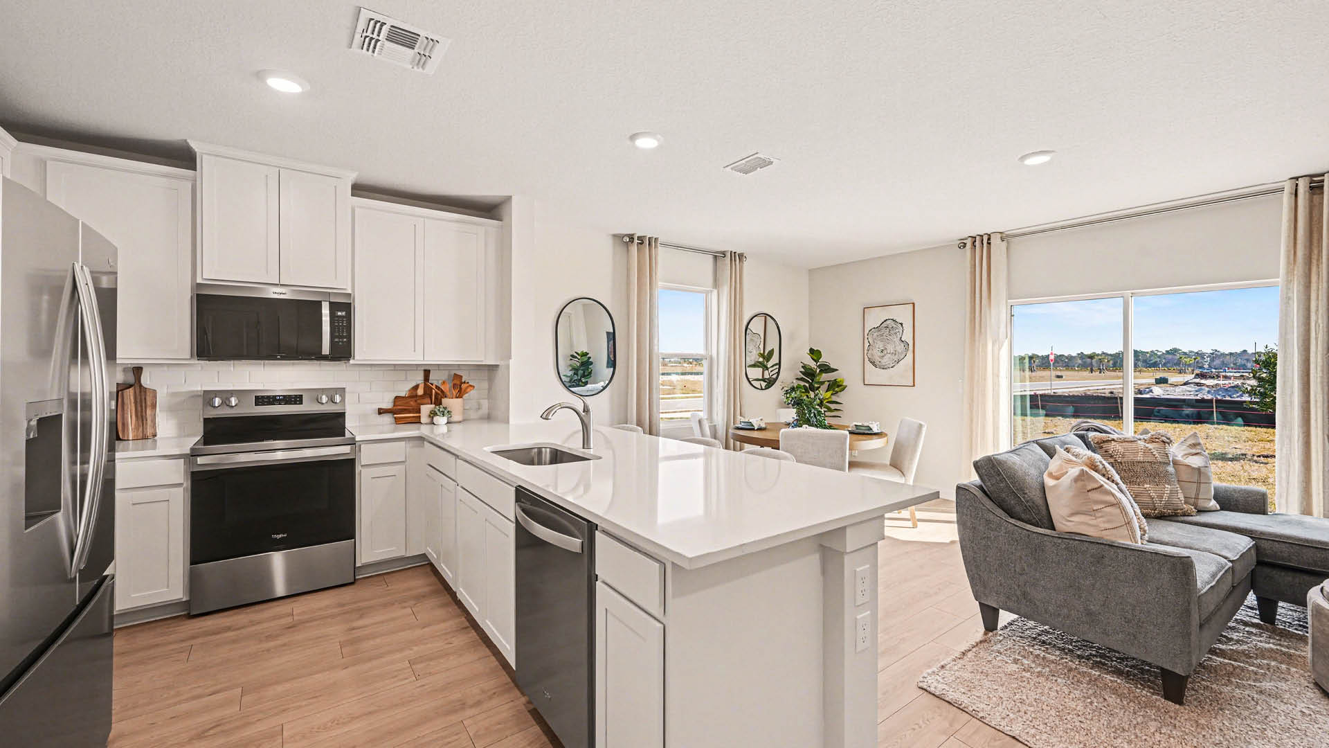 A large kitchen with a peninsula with seating and stainless steel appliances. The dining room and living room are visible beyond the island and illuminated by natural light from the window and sliding glass door.