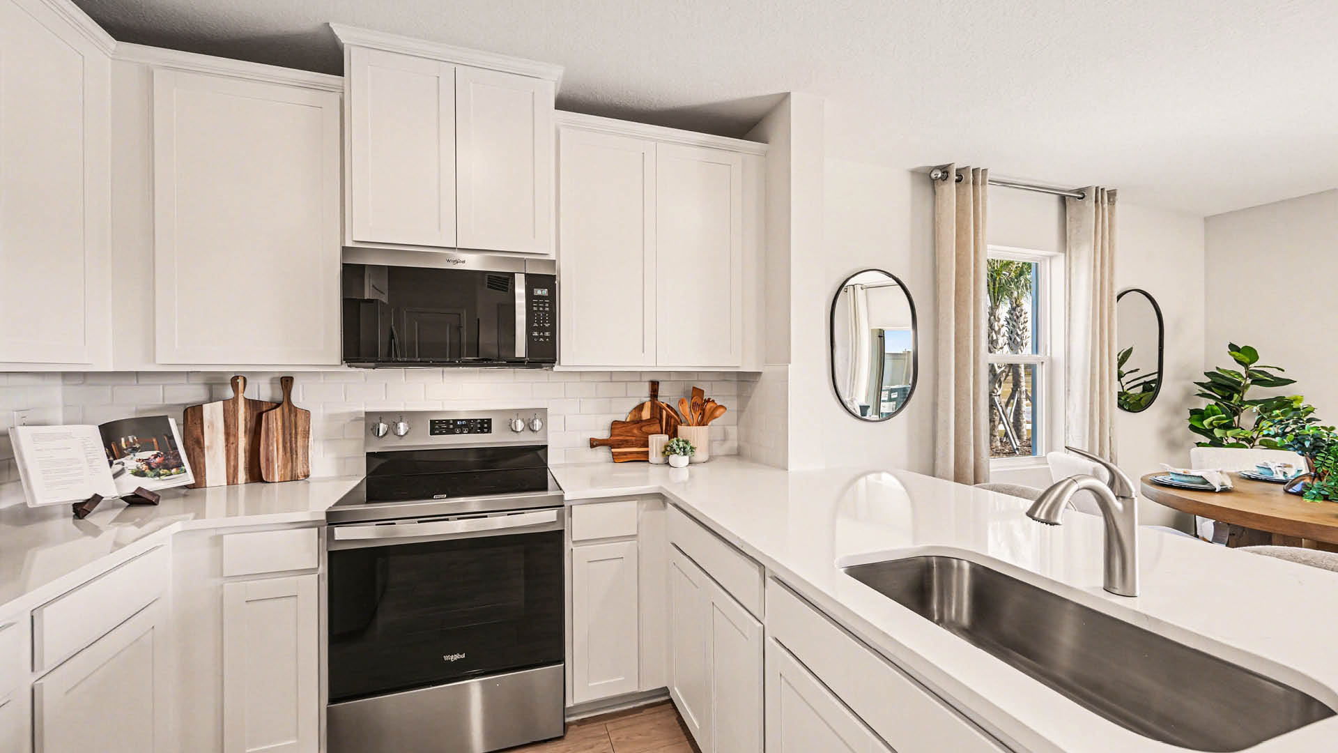 A large kitchen with quartz countertops, stainless steel appliances, and modern, white cabinetry. A window and dining area are visible.