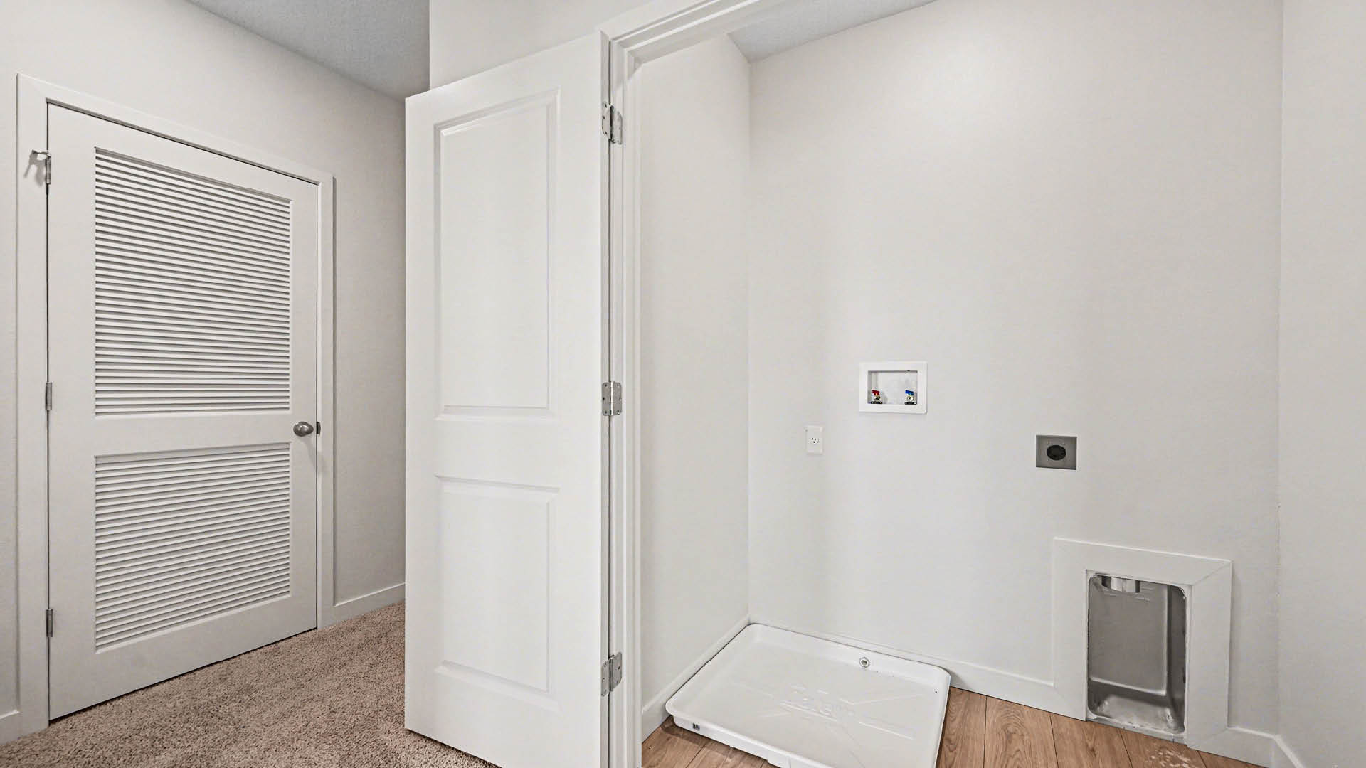 A small laundry room with light-colored walls, plank flooring, and utility connections on one wall, featuring a closed door on the left.