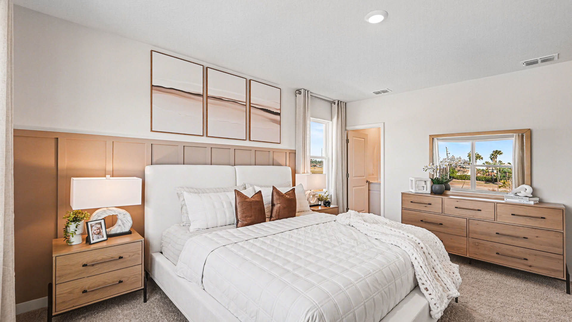 A large bedroom with beige carpet, white walls, and a window, allowing natural light to illuminate the space. An open door leading to an ensuite bathroom is visible.