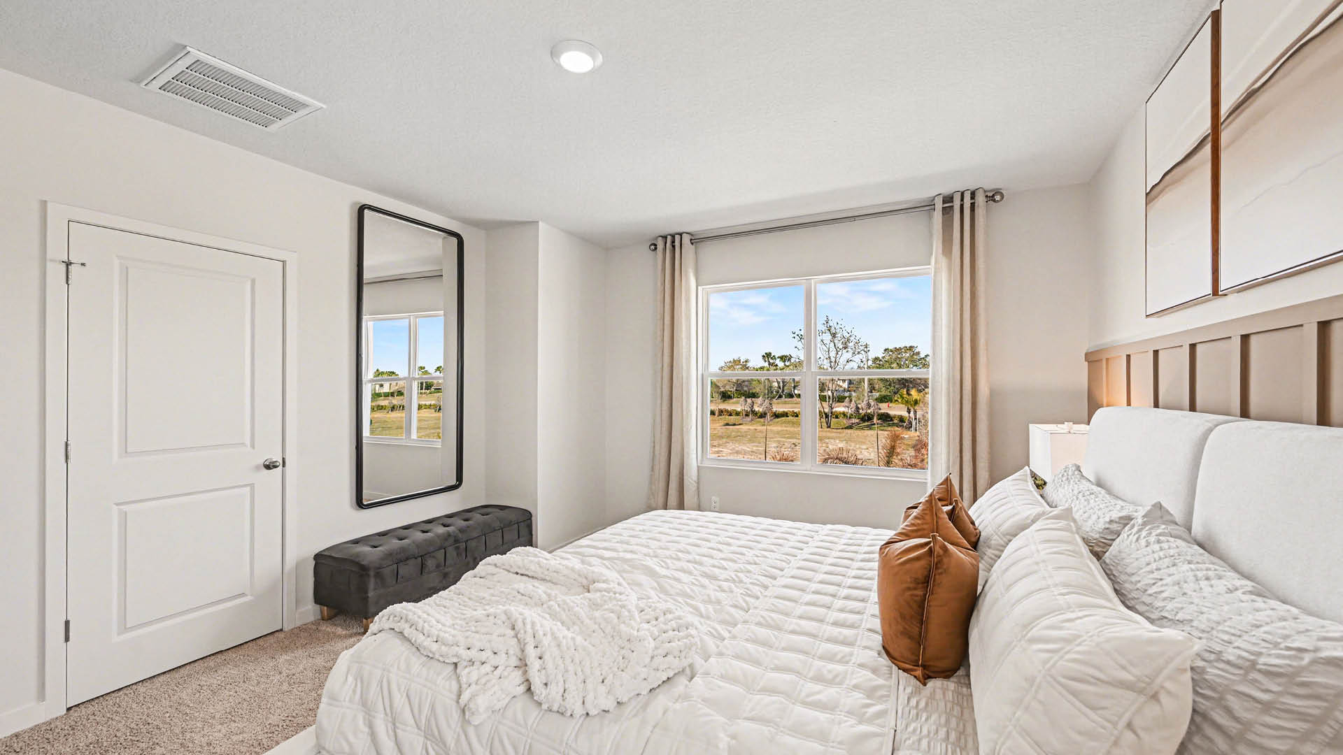 A large bedroom with beige carpet, white walls, and a window, allowing natural light to illuminate the space. A closed door is visible.