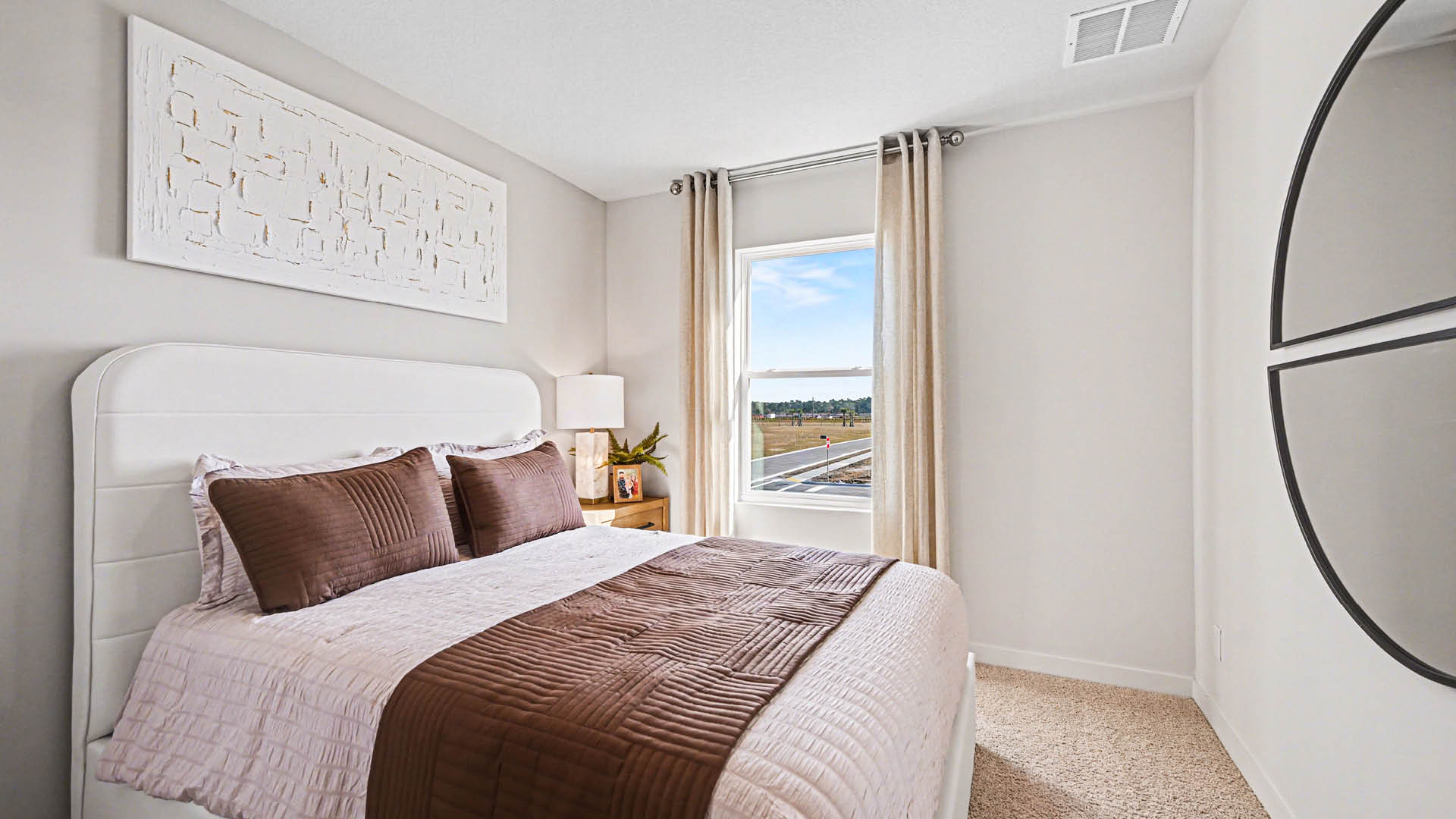 A bedroom with a bed, a window, and a painting on the wall. The room has white walls and beige carpet.