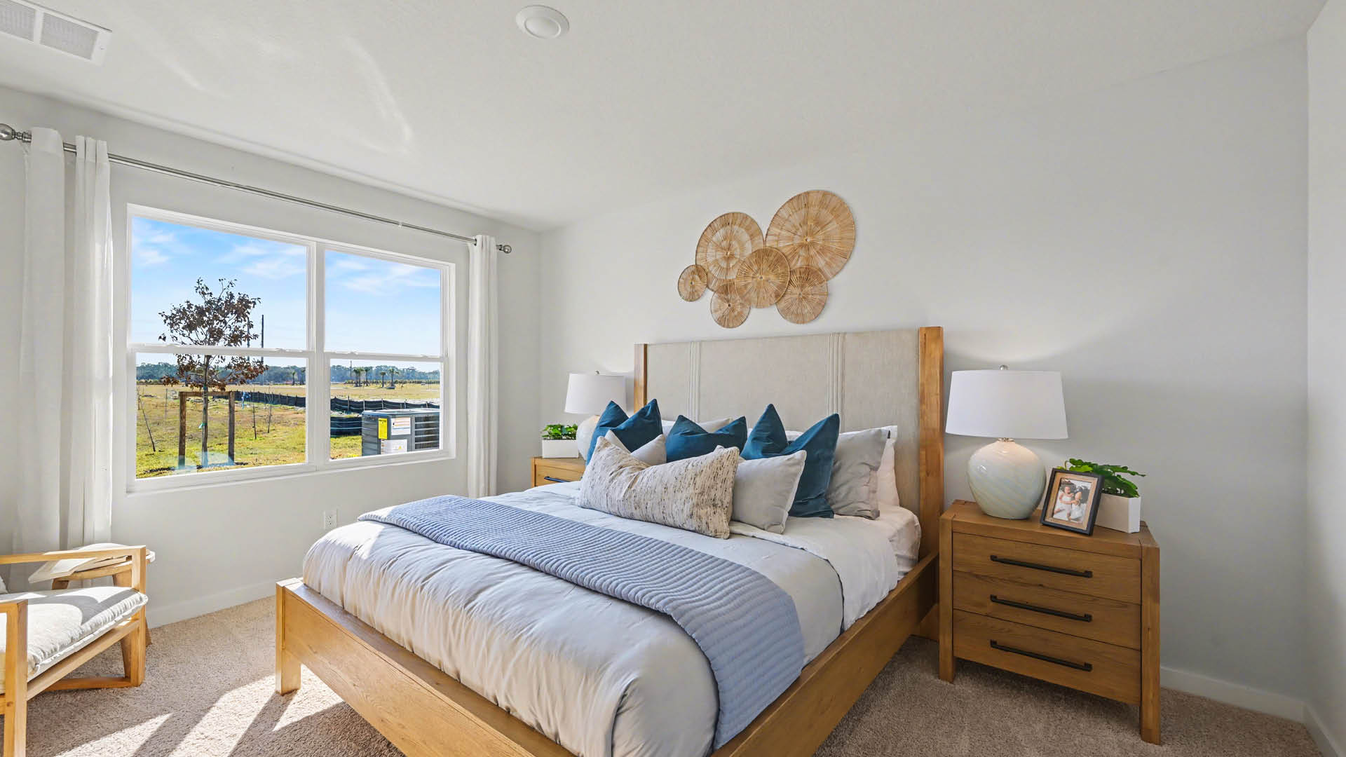 A bright bedroom with a large window where natural light is streaming in. The room has beige carpet, white walls, and a bed.